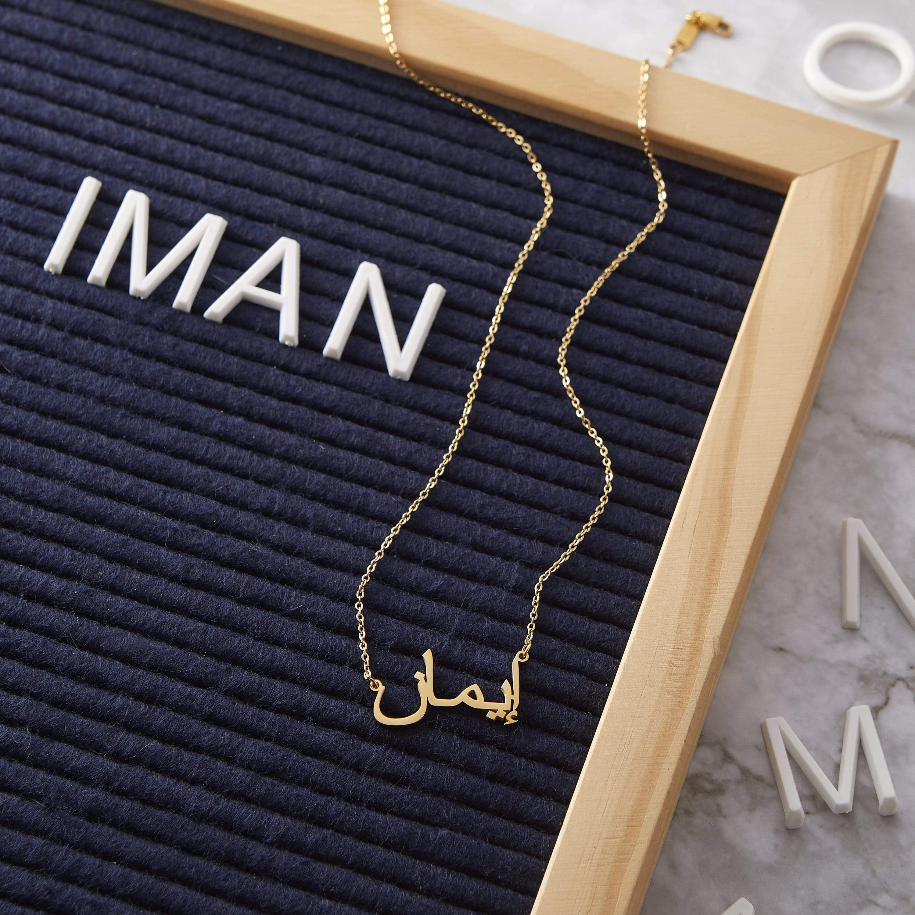 A gold necklace with a pendant that spells out the name "Iman" in Arabic script. The necklace is displayed on a textured dark blue surface, with the name "IMAN" spelled out in large white block letters in the background. A wooden frame surrounds the display.