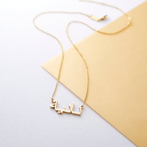 A gold necklace with a name pendant in Arabic script. The chain is delicate and gold. The necklace is displayed on a light beige background.