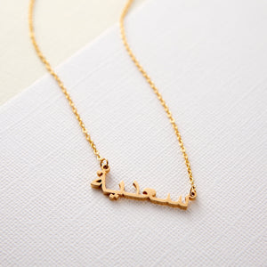A gold necklace with a name pendant in Arabic script. The chain is delicate and gold. The necklace is displayed on a light beige background.