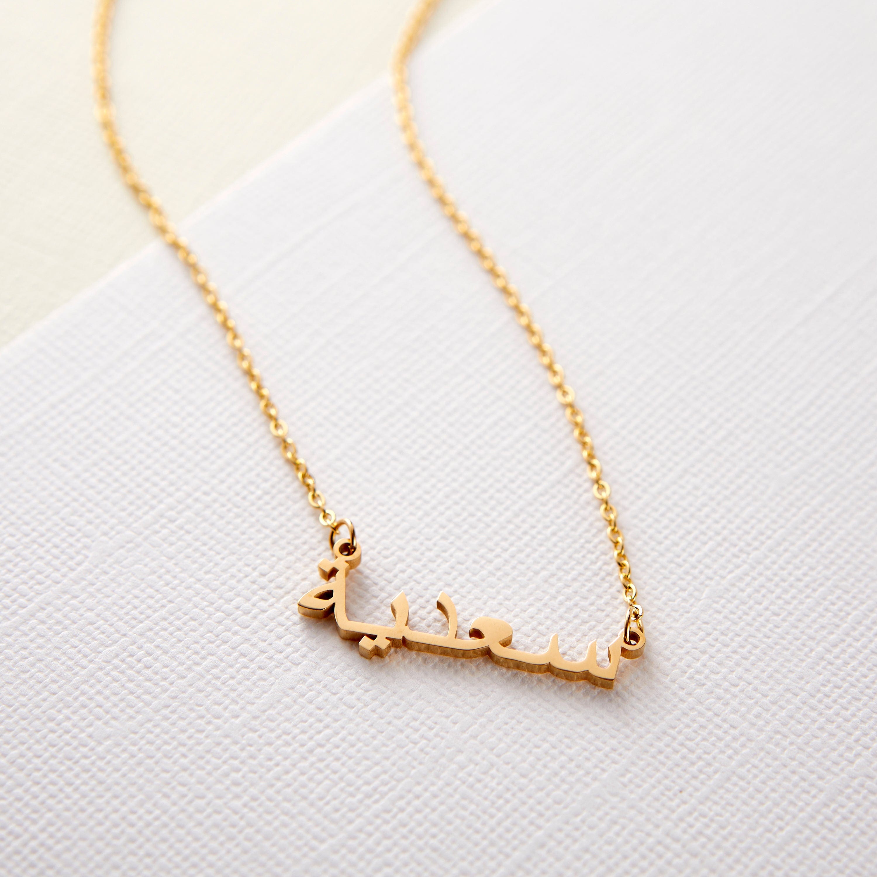 A gold necklace with a name pendant in Arabic script. The chain is delicate and gold. The necklace is displayed on a light beige background.