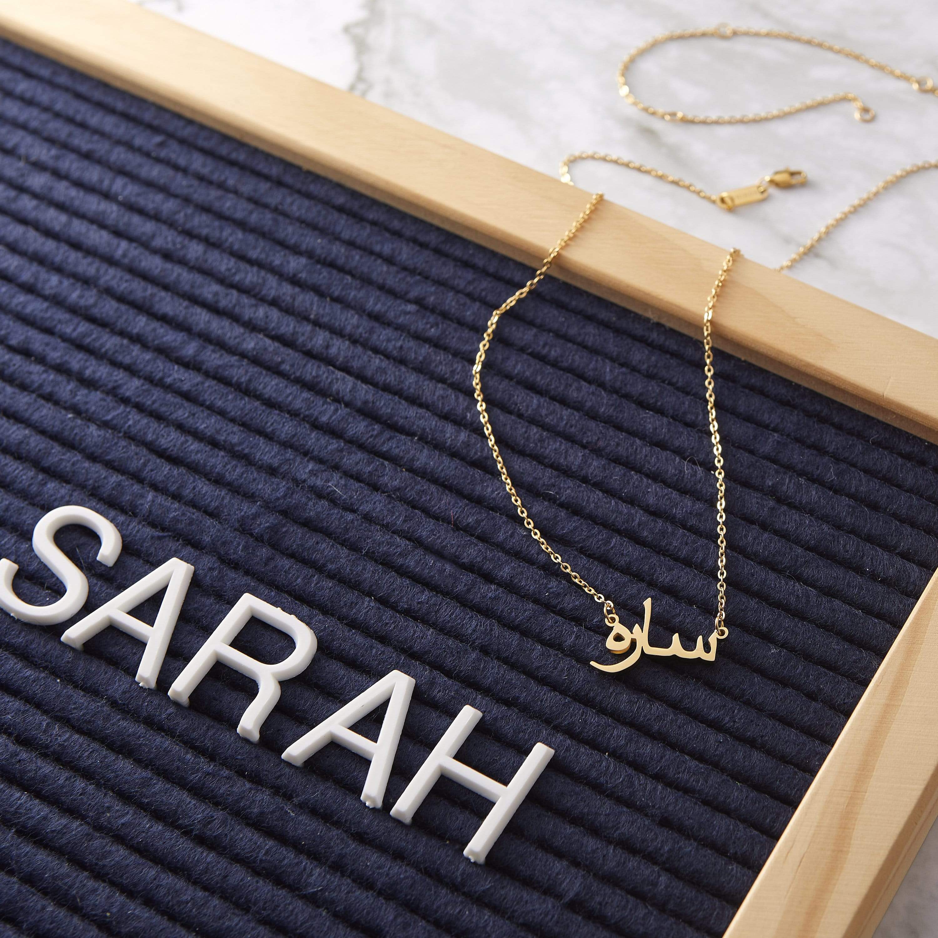 A gold necklace with a pendant that spells out the name "Iman" in Arabic script. The necklace is displayed on a textured dark blue surface, with the name "SARAH" spelled out in large white block letters in the background. A wooden frame surrounds the display.