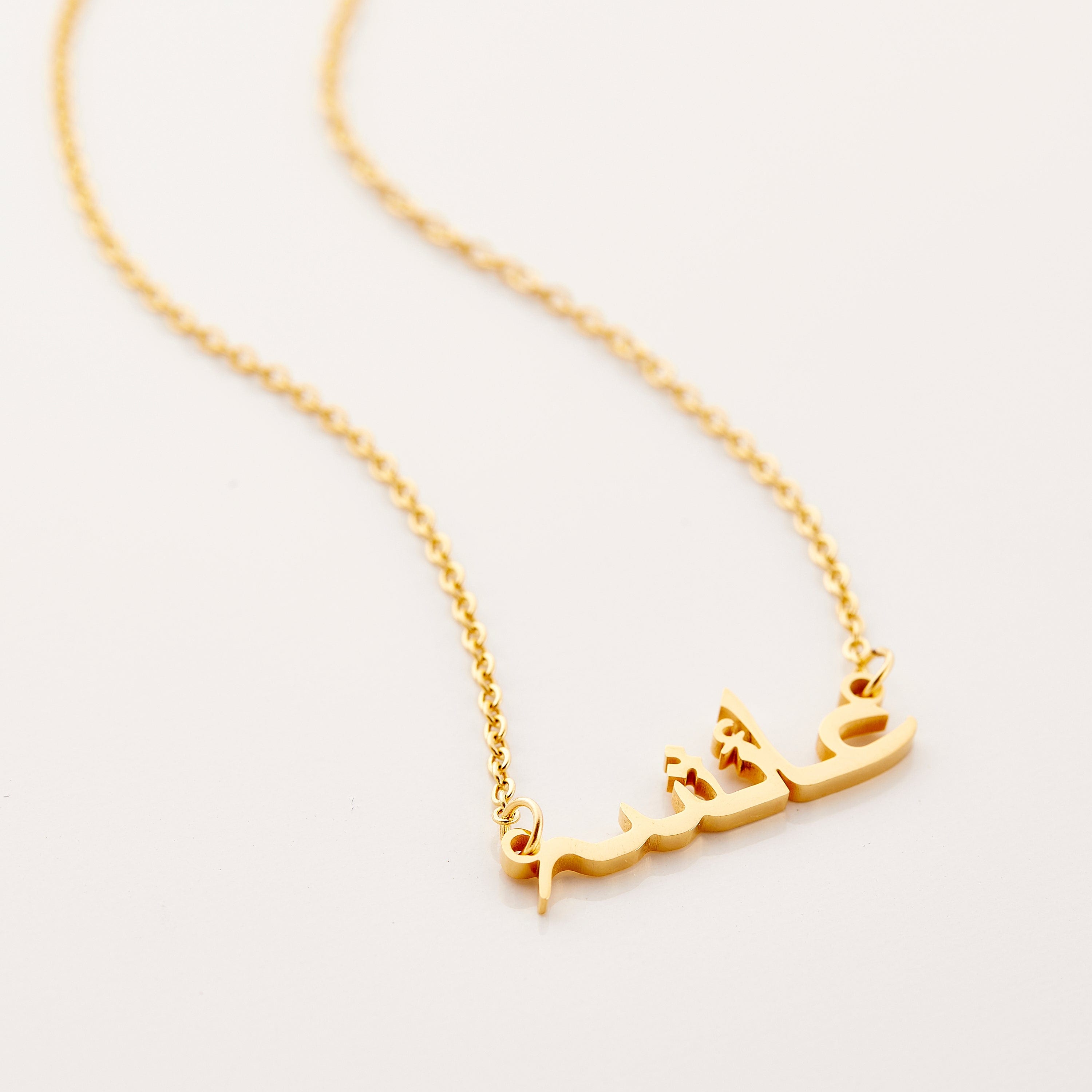 A gold necklace with a name pendant in Arabic script. The chain is delicate and gold. The necklace is displayed on a light beige background.