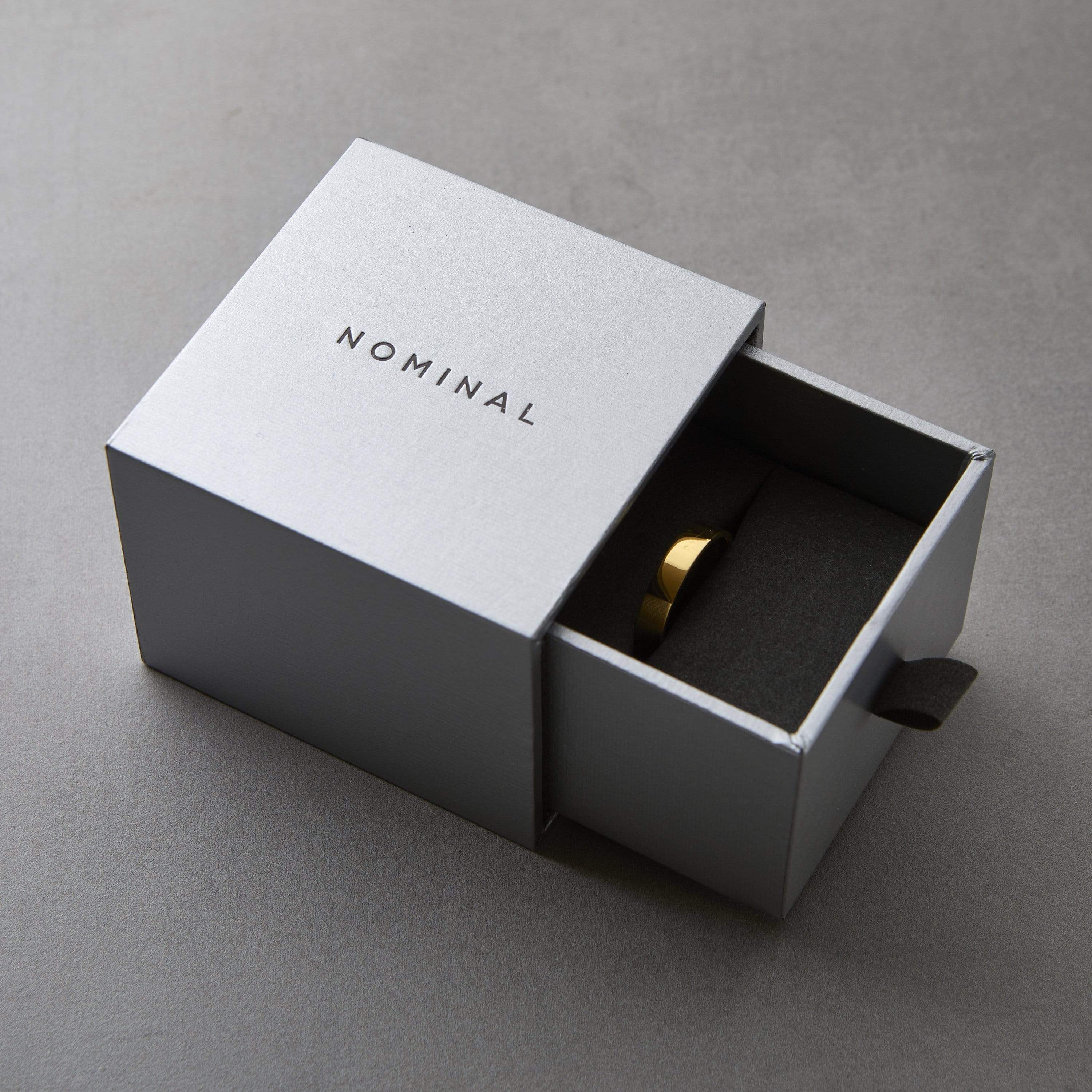 Silver Nominal drawer-style gift box with black text “NOMINAL” on the lid, partially opened to reveal a gold ring inside, resting in black foam lining against a gray background.