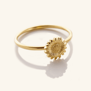 Close-up of a thin gold ring featuring a detailed sunflower design on top, photographed against a light background.