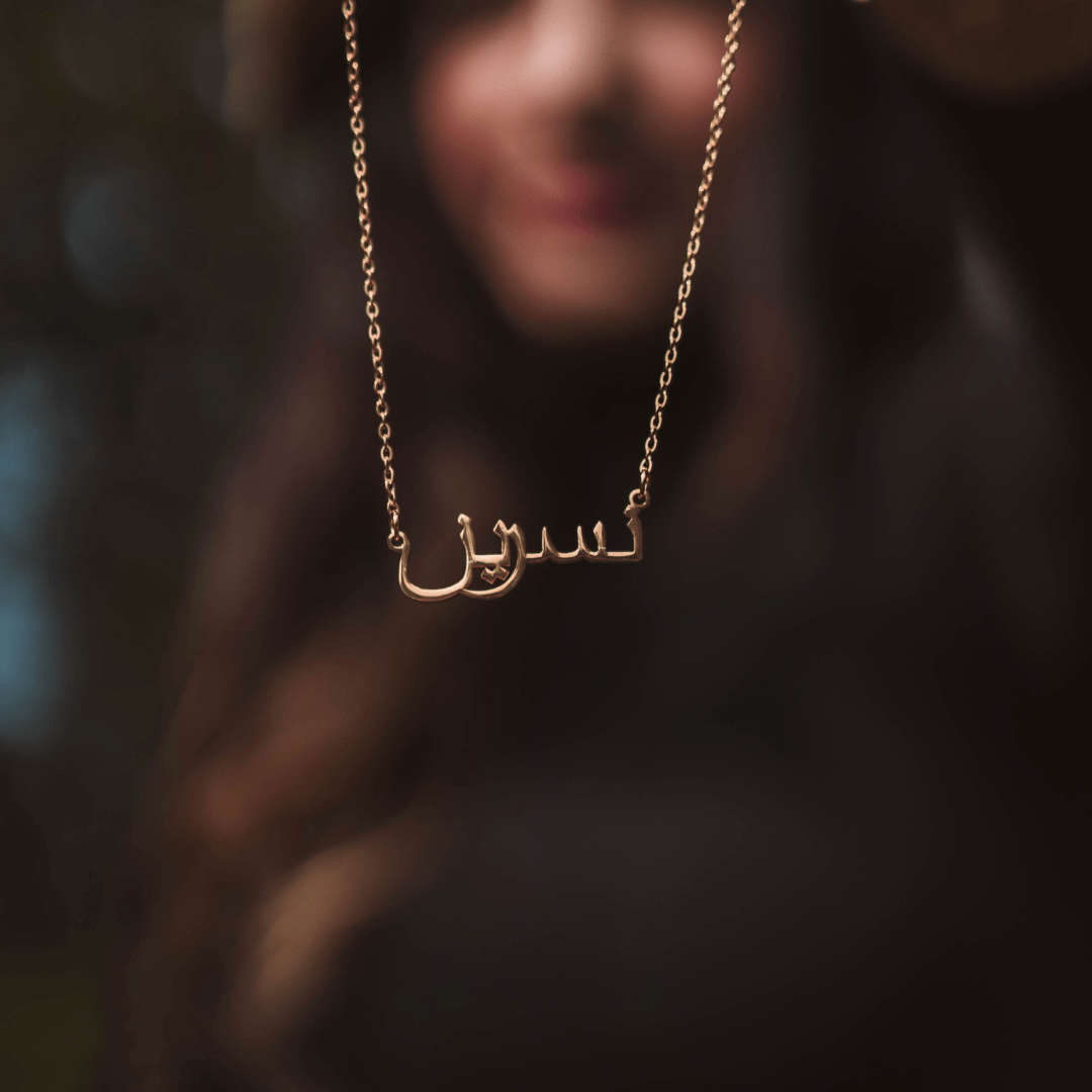 A gold necklace with a pendant displaying a name written in elegant Arabic script. The necklace hangs in focus against a blurred background of a person with long, dark hair and a soft smile. The lighting highlights the gold of the necklace and the intricate details of the Arabic lettering.