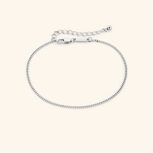 Slim silver curb chain anklet with lobster clasp and extender, featuring a small tag engraved with "NOMINAL," laid out on a clean beige surface.