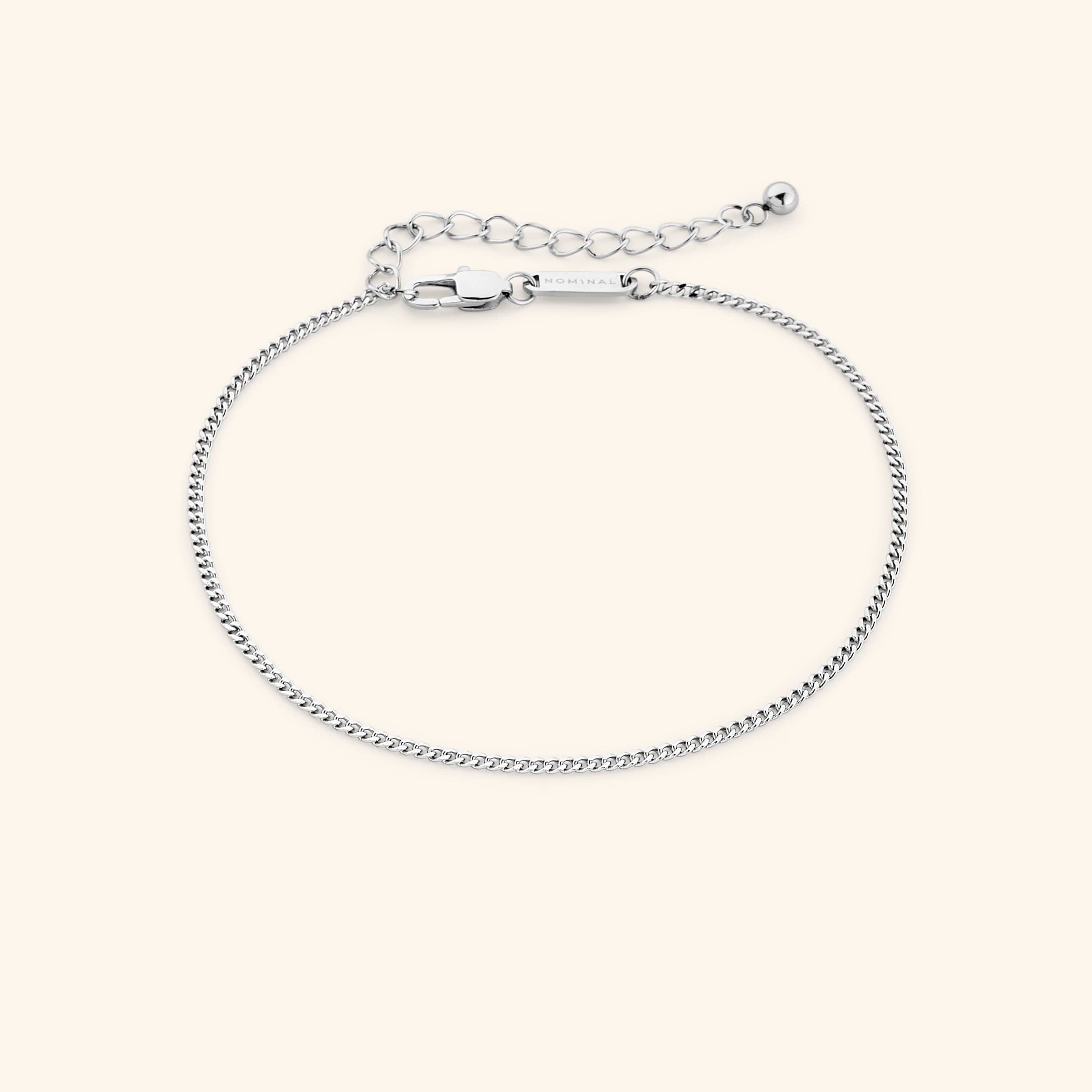 Slim silver curb chain anklet with lobster clasp and extender, featuring a small tag engraved with "NOMINAL," laid out on a clean beige surface.
