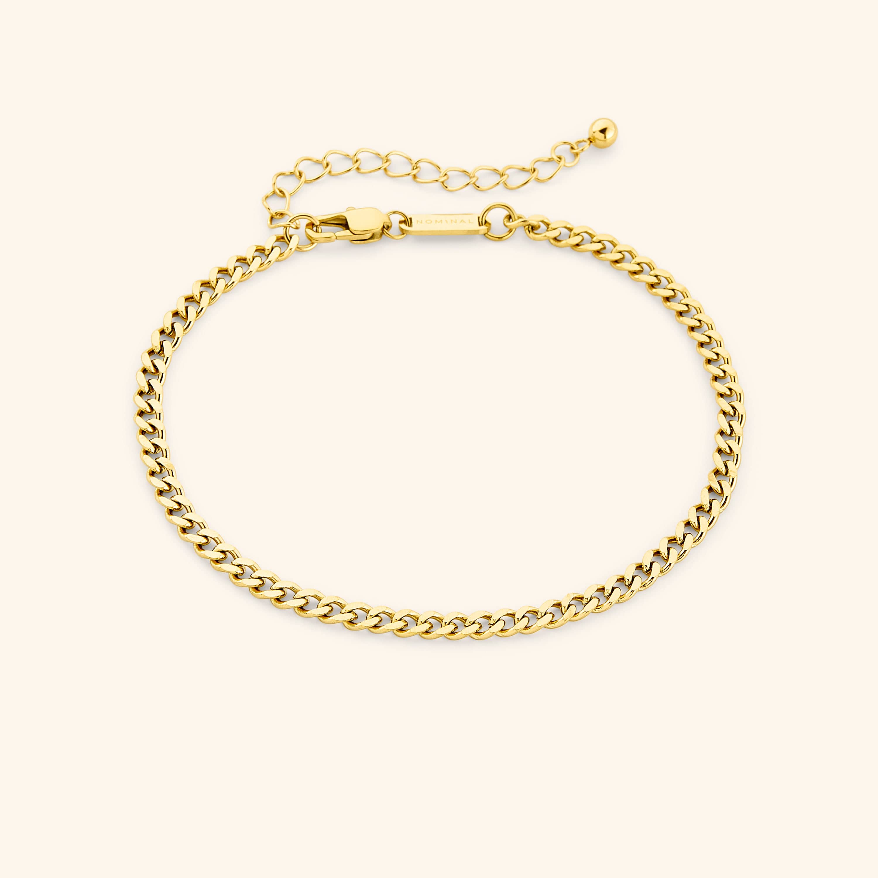 Chunky gold curb chain anklet with lobster clasp and extender, tag engraved with "NOMINAL" visible near the clasp, laid out on a soft beige surface.