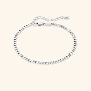 Thick silver curb chain anklet with extender and lobster clasp, rectangular tag engraved with "NOMINAL" near the clasp, styled on a soft beige background.