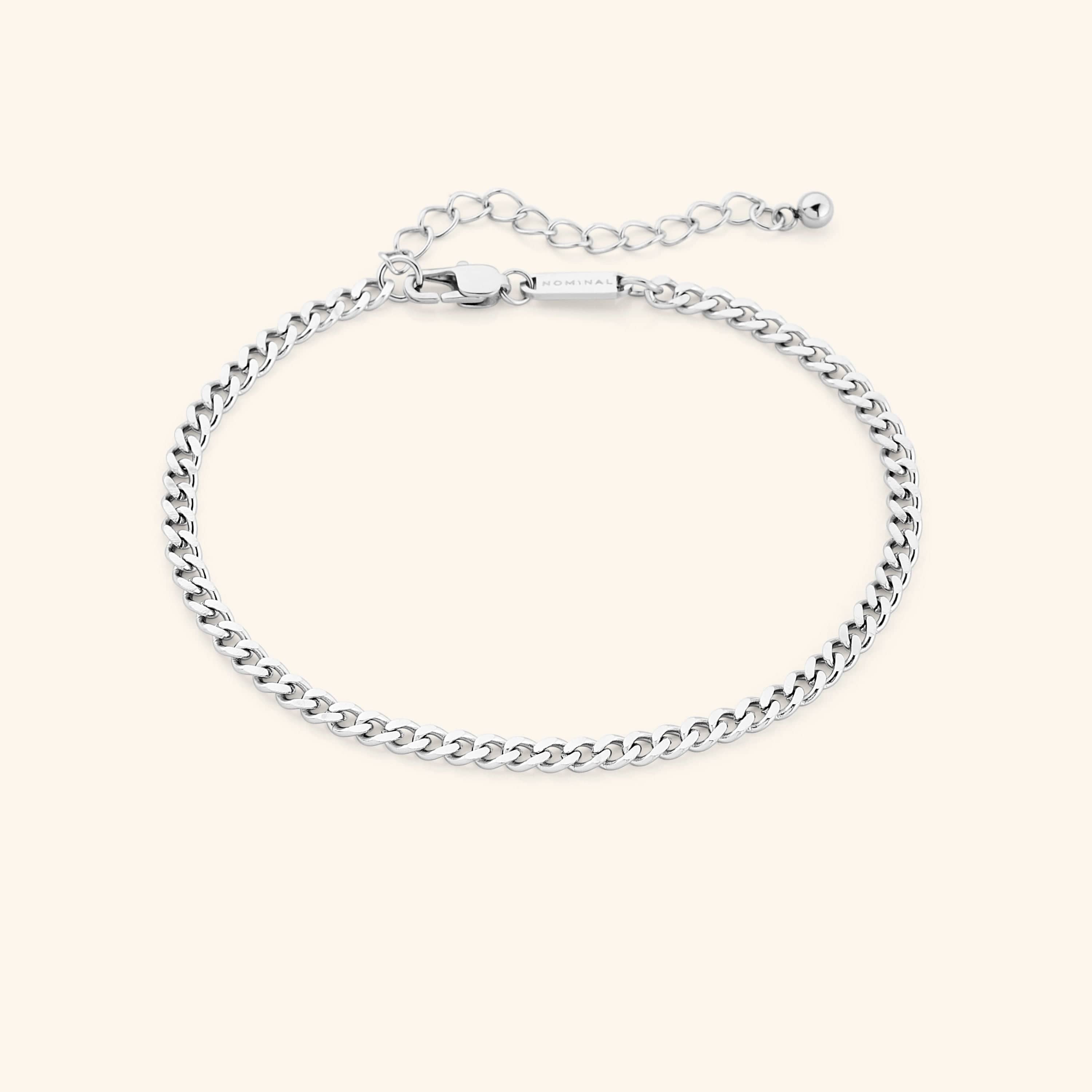 Thick silver curb chain anklet with extender and lobster clasp, rectangular tag engraved with "NOMINAL" near the clasp, styled on a soft beige background.