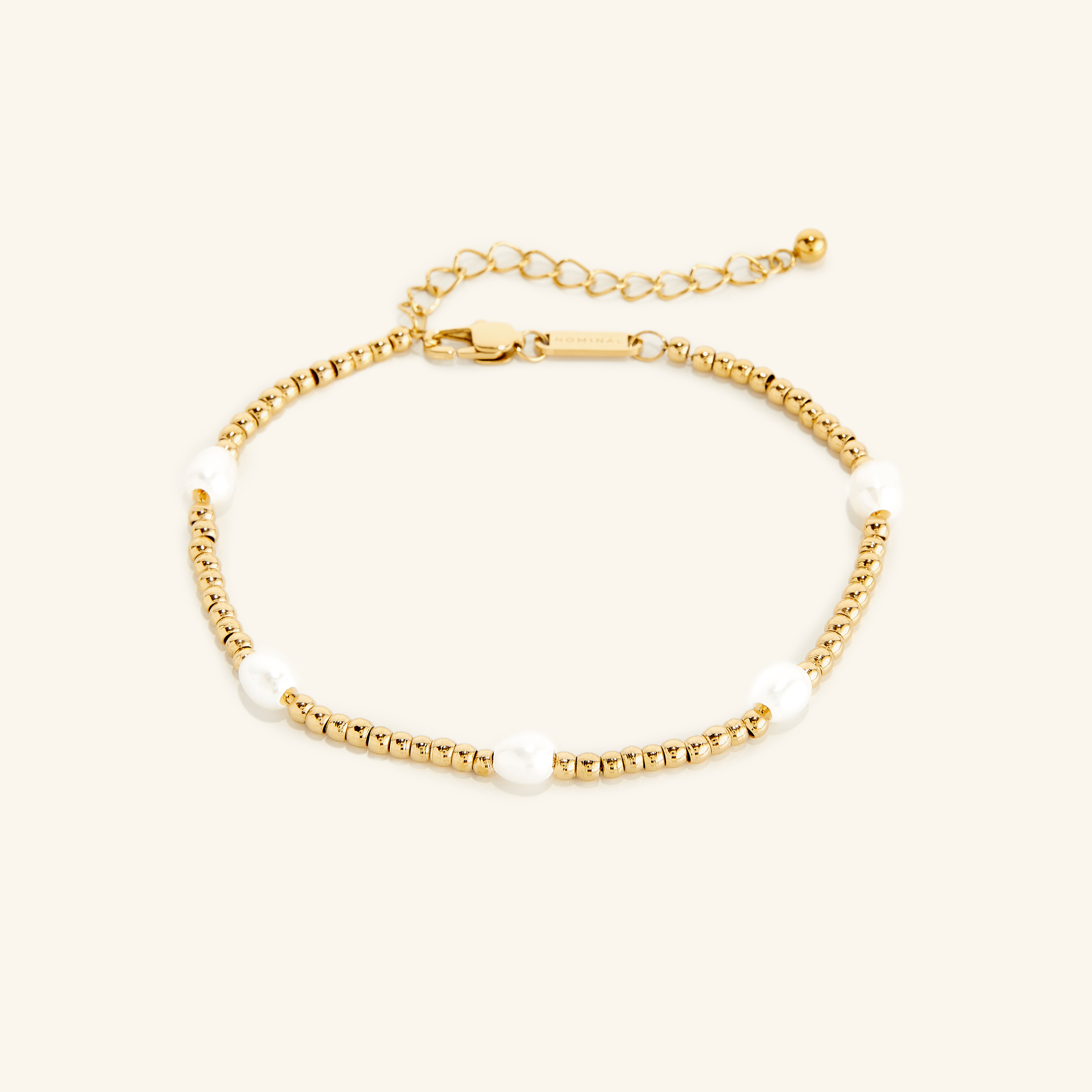 Gold anklet with small round beads and five evenly spaced white pearls, shown on a cream background with an adjustable clasp and extender chain engraved with 'NOMINAL'.