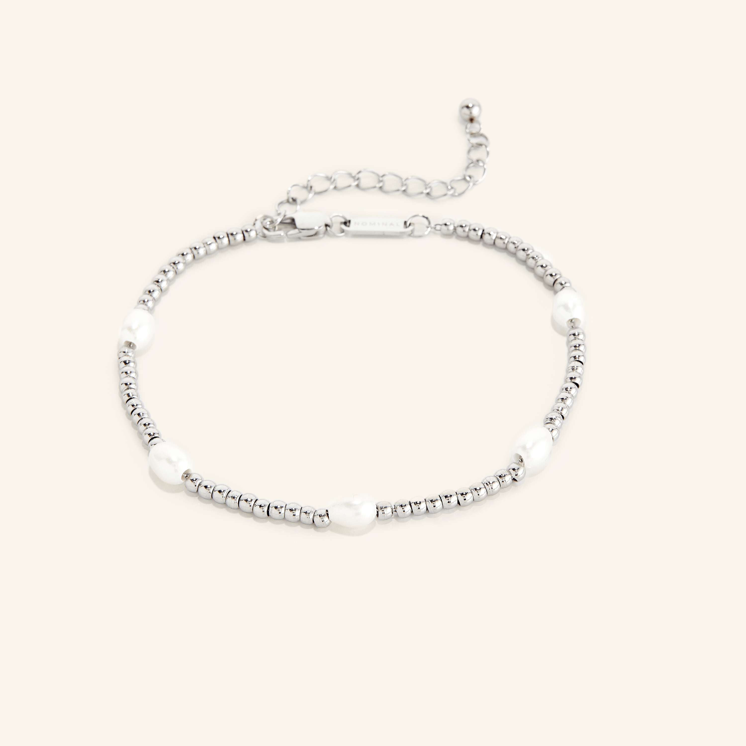 Silver anklet with small round beads and five evenly spaced white pearls, shown on a cream background with an adjustable clasp and extender chain engraved with 'NOMINAL'.