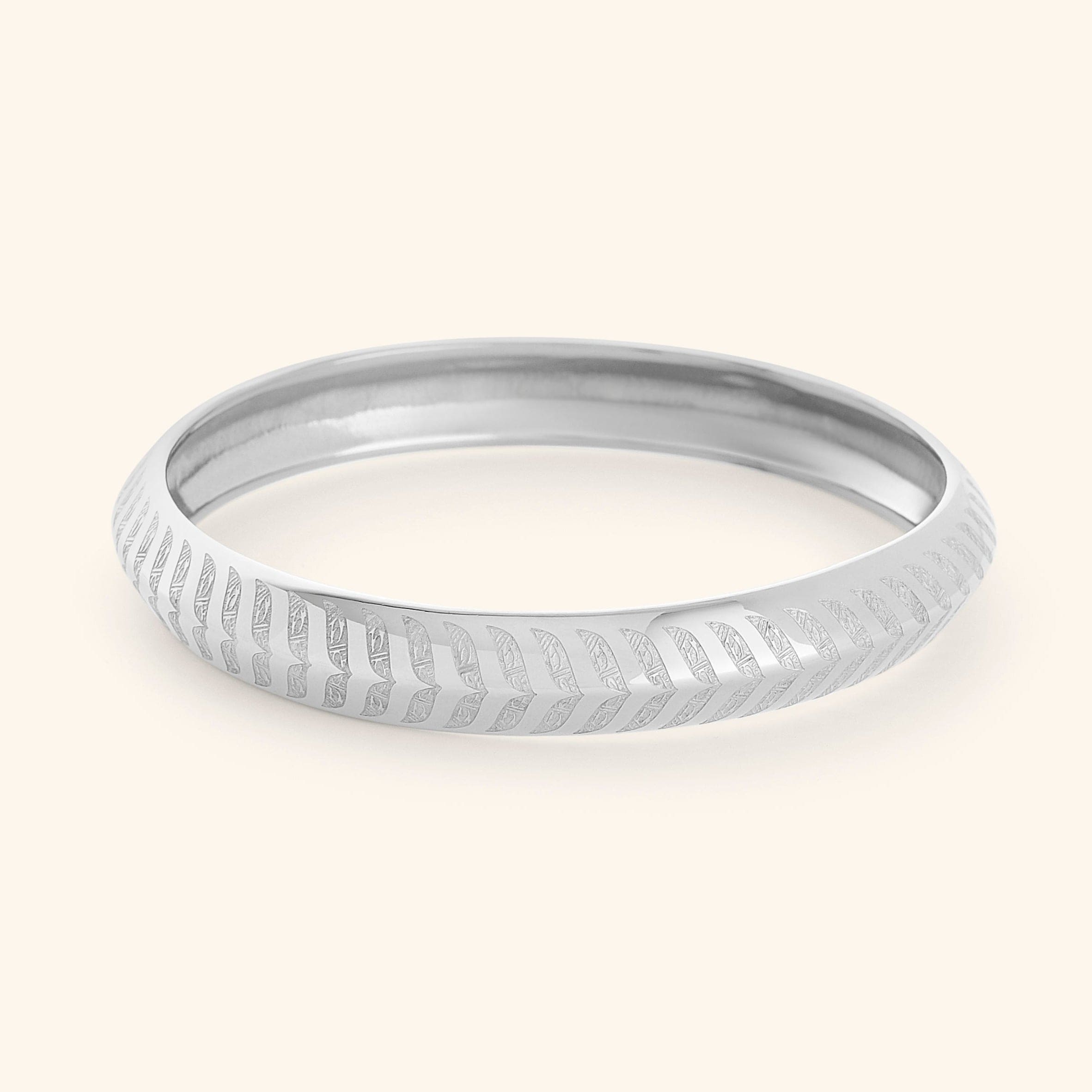 A silver "Chevron Bangle" designed for women as part of the Aadil x Nominal collection by Nominal. The bangle has a smooth, rounded surface and is decorated with intricate, stylized chevron or feather-like carvings that run around its entire circumference.