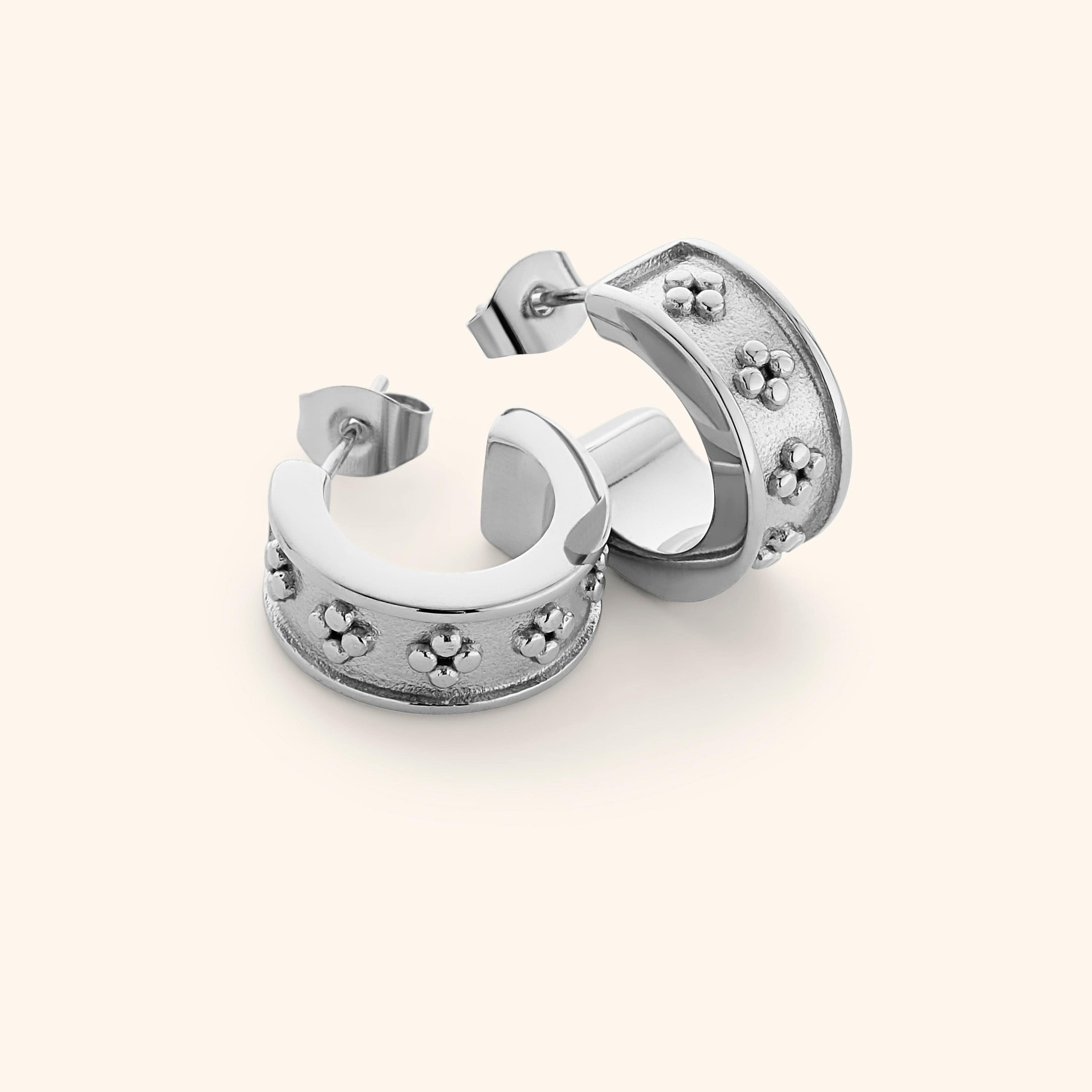 Flat lay of silver Henna Hoops, wide curved band with raised dotted floral motifs on a textured background.