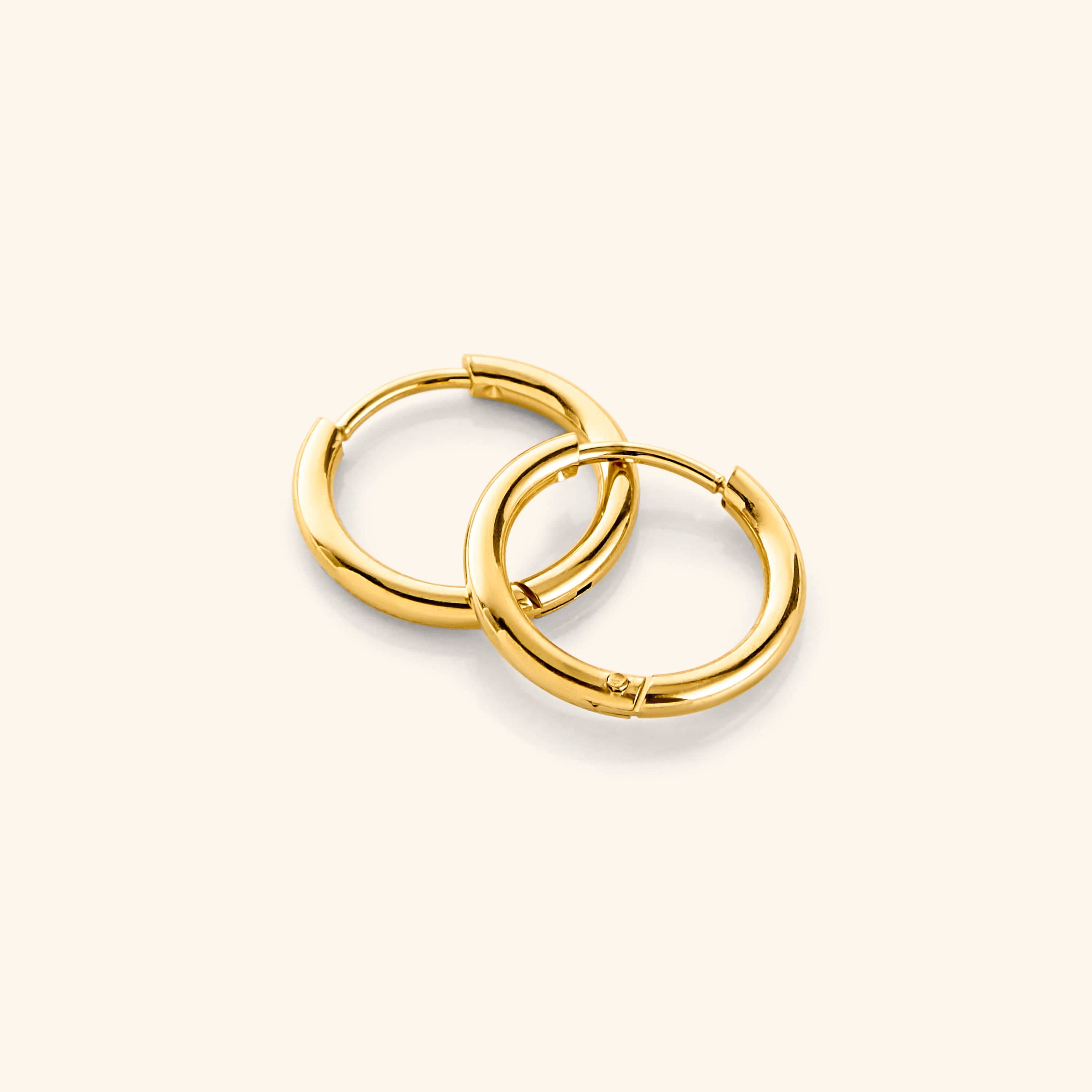 a pair of 18k gold plated Huggie Essential Hoops | 12mm against a light background.