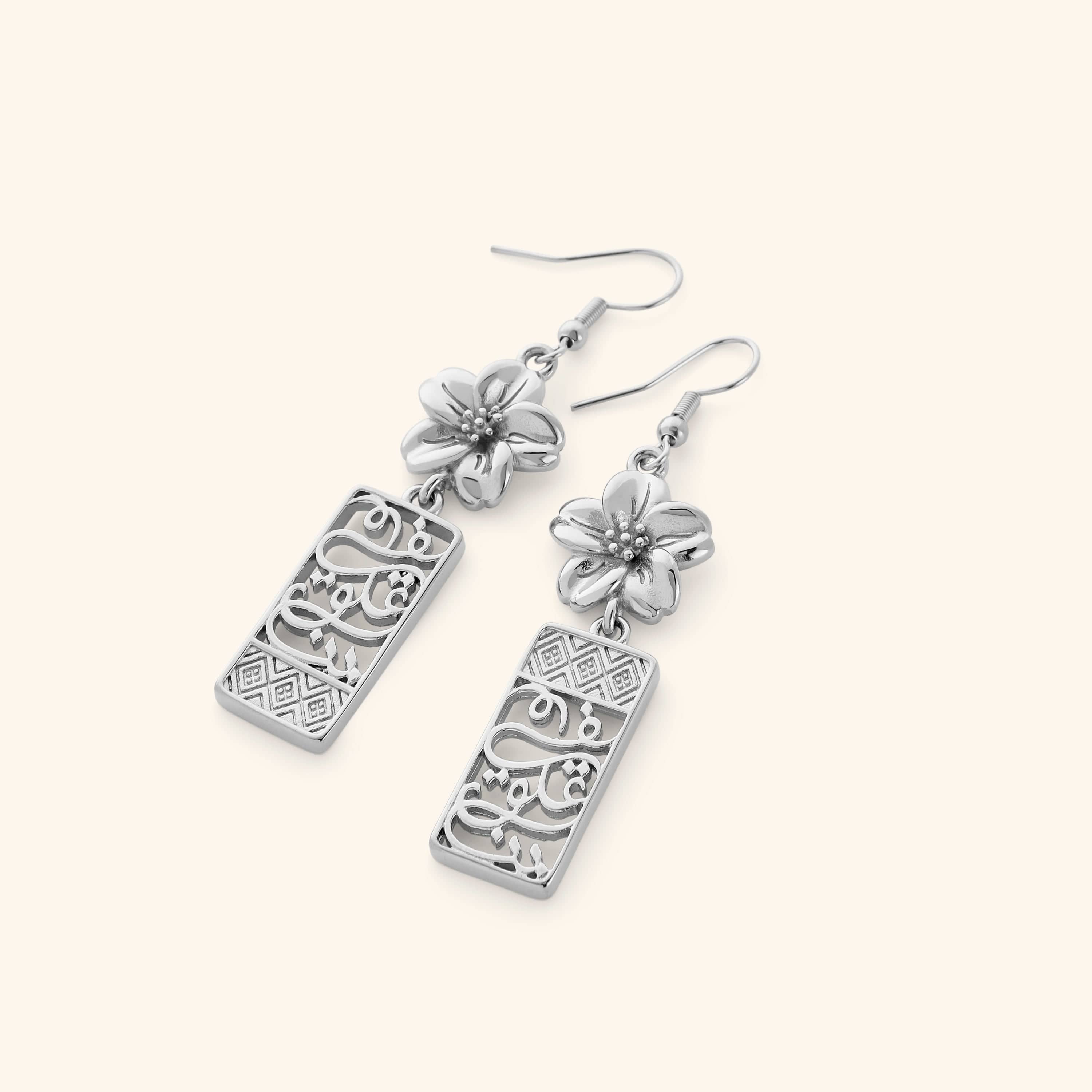 A pair of silver dangling earrings featuring patterns found in the Damascus masjids and courtyards and Arabic calligraphy "in our hearts" design finished with blooming petals of a Jasmine at the top.