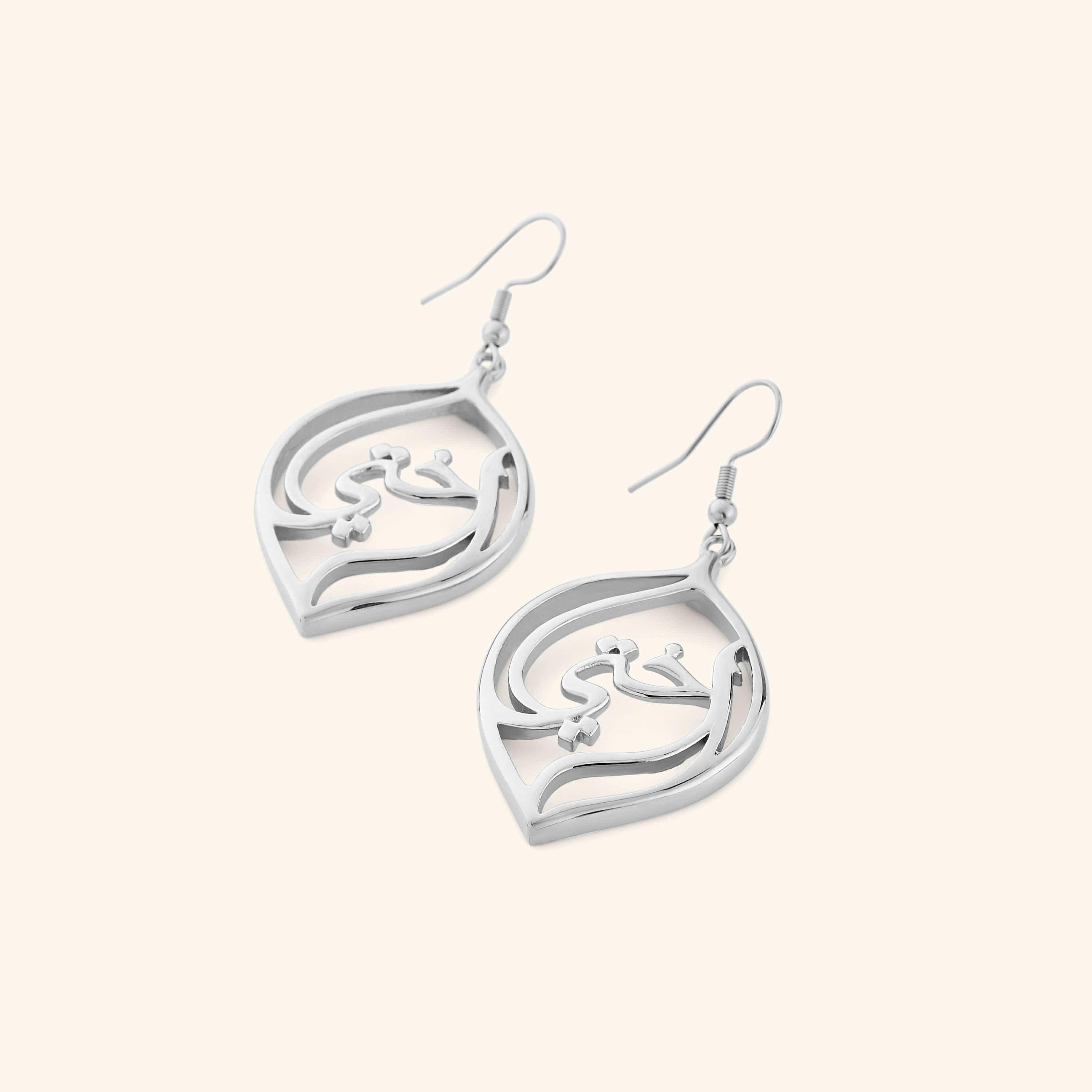 A pair of silver earrings with a droplet framing the Arabic word for "my sister", أختي (ukhti).