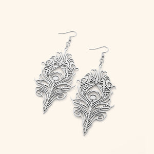 The image shows a pair of silver "Peacock Feather Earrings" from the Aadil x Nominal collection. The earrings are intricately designed in the shape of peacock feathers, with detailed cut-out patterns that mimic the barbs and eye of the feather. This appears to be a product shot showcasing the earrings' design and craftsmanship.