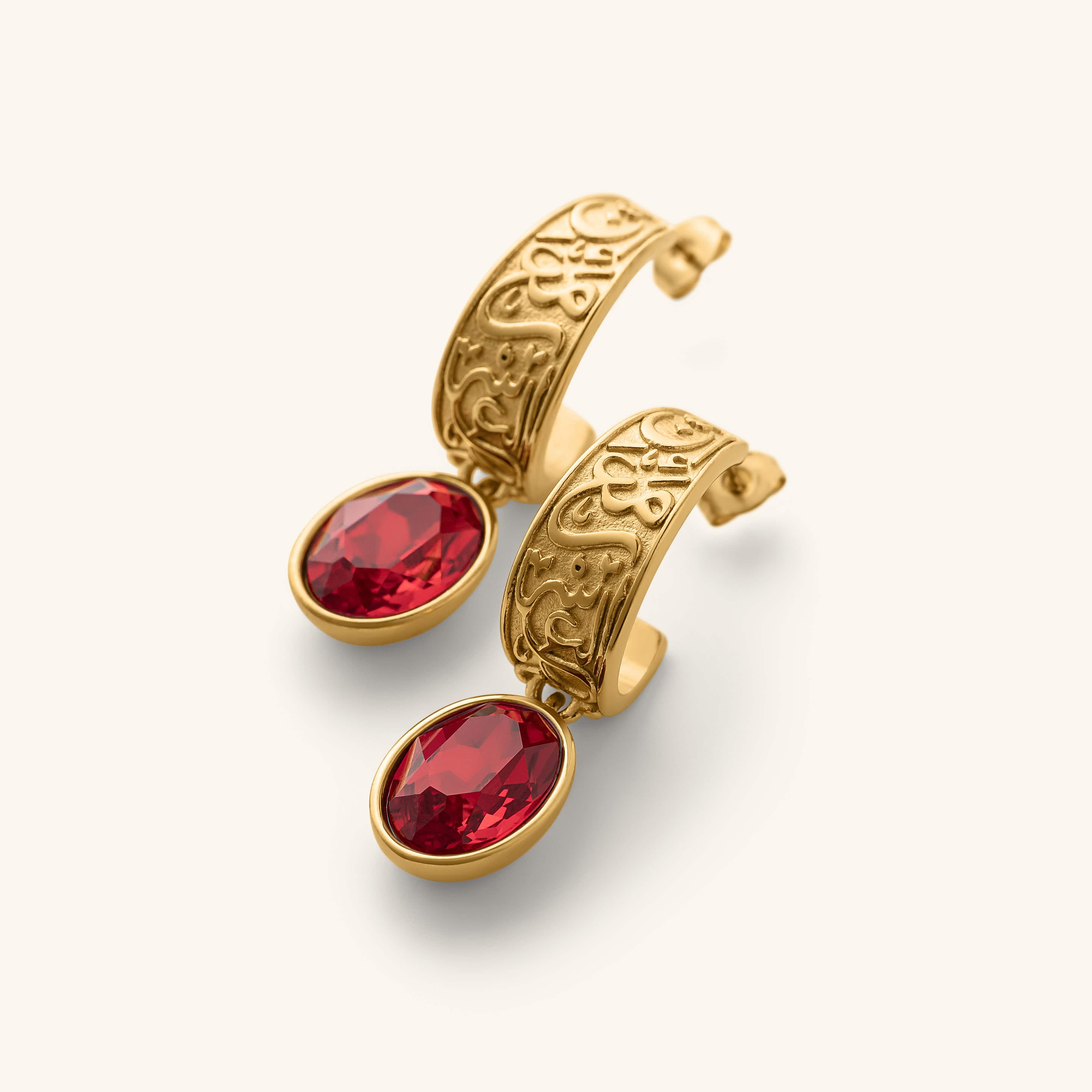"With Hardship Comes Ease” Earrings in gold, featuring engraved Arabic calligraphy along the curved band with an oval red gemstone drop hanging below, styled against a neutral background.
