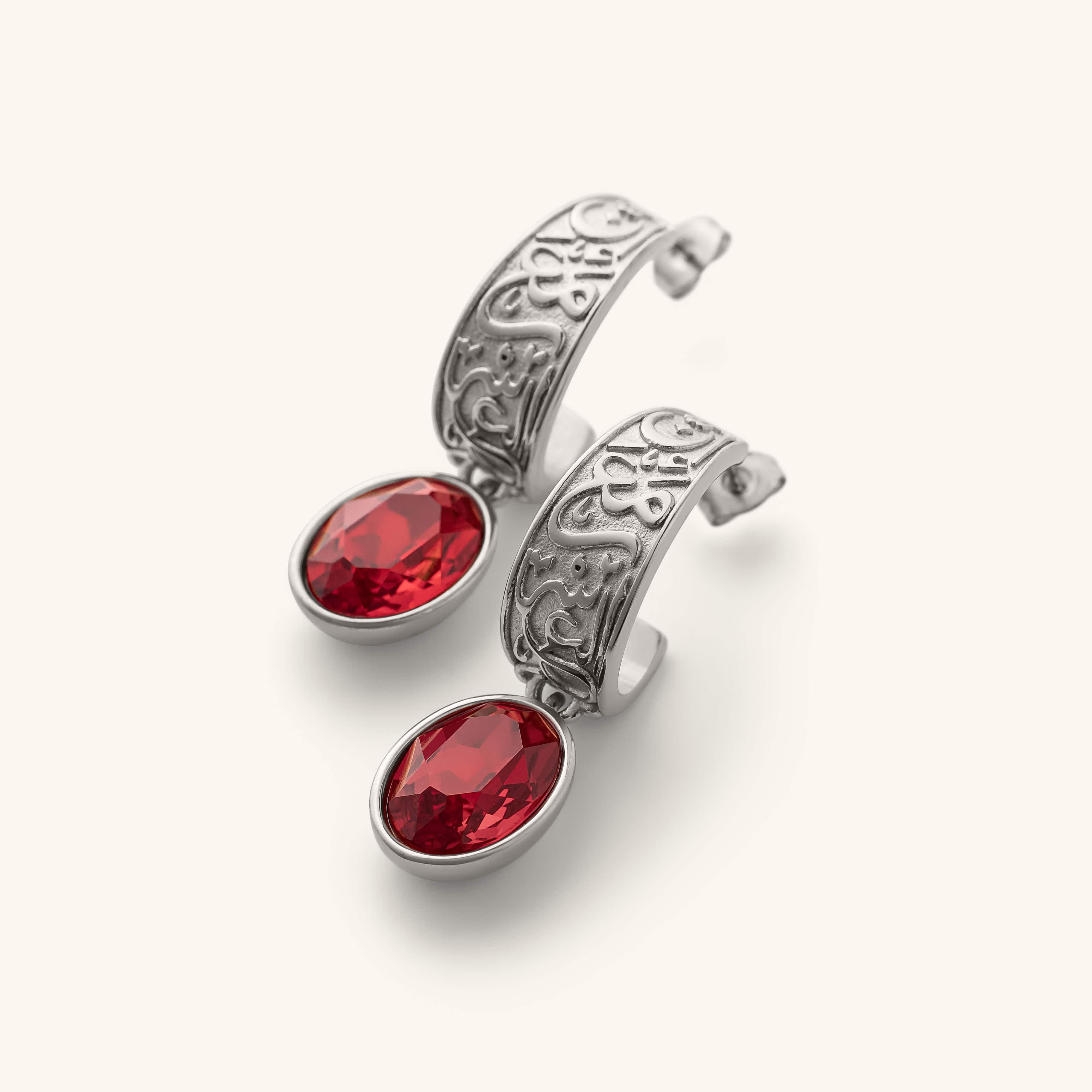 "With Hardship Comes Ease” Earrings in silver, featuring engraved Arabic calligraphy along the curved band with an oval red gemstone drop hanging below, styled against a neutral background.