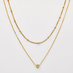 Gold necklace set featuring a bead-accented chain layered with a solitaire bezel-set pendant necklace.