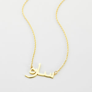 A close-up of the Custom Name Necklace in 14K solid gold, featuring the Arabic name "ساره" ("Sarah") in elegant calligraphy. The pendant is suspended from a delicate gold chain, highlighting the craftsmanship and personalized design.