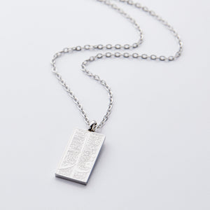 The image shows a delicate gold necklace displayed against a clean, light gray background. The chain is thin and elegant, forming a gentle curve that draws the eye toward the pendant hanging at the center.  The pendant is rectangular and made of silver, with a smooth, polished finish that reflects light subtly. Its surface features intricate engraved patterns that add texture and visual interest.