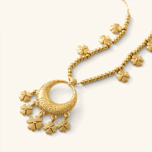 Close-up of the Levantine Garden Necklace, featuring a gold pendant engraved with floral Levantine patterns and adorned with multiple dangling clover-shaped charms. The pendant hangs from a beaded gold chain with additional clover charms spaced along it.