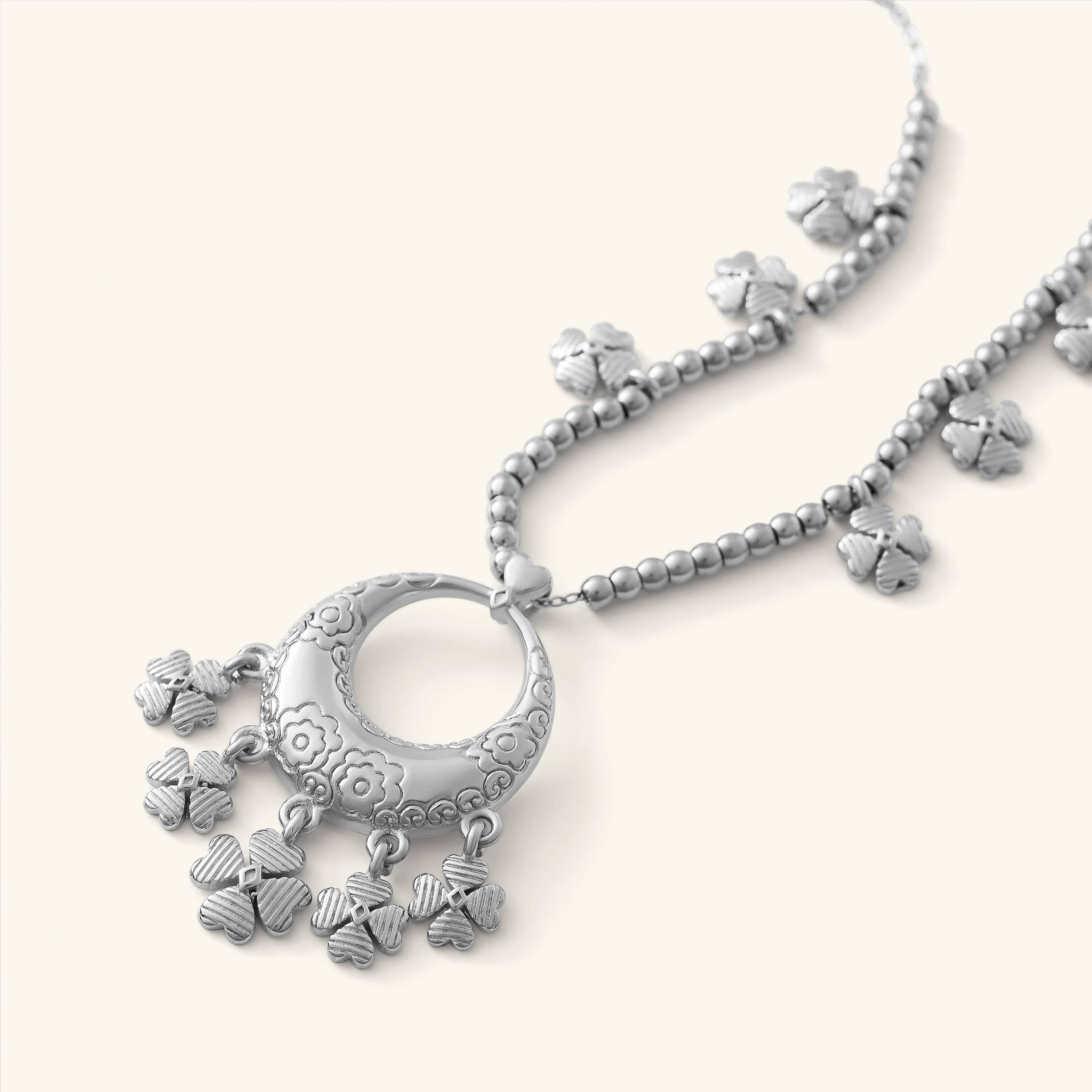 Close-up of the Levantine Garden Necklace, featuring a silver pendant engraved with floral Levantine patterns and adorned with multiple dangling clover-shaped charms. The pendant hangs from a beaded gold chain with additional clover charms spaced along it.
