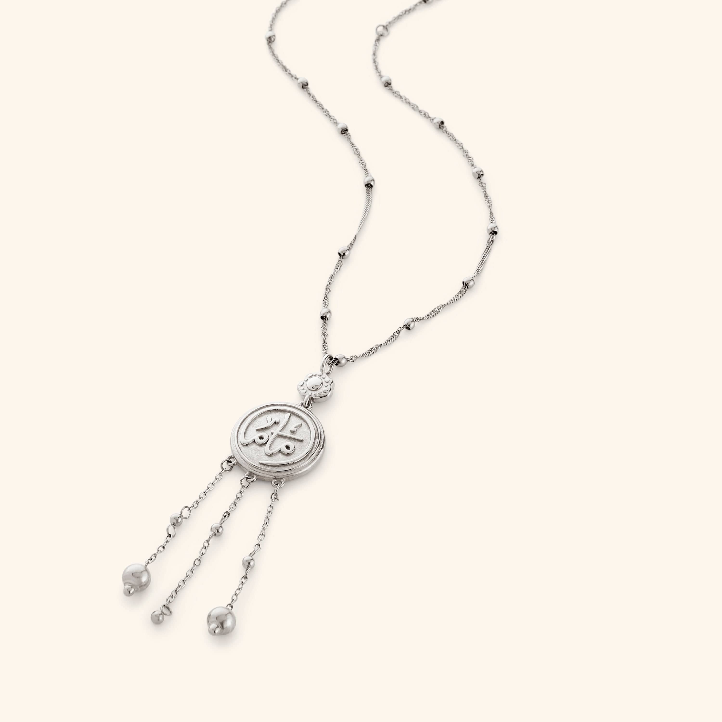 Silver necklace that features a coin pendant stamped with ماما (Mama) in Arabic, suspended from a beaded chain with three strands falling beneath, stamped with a miniature floral at the top.