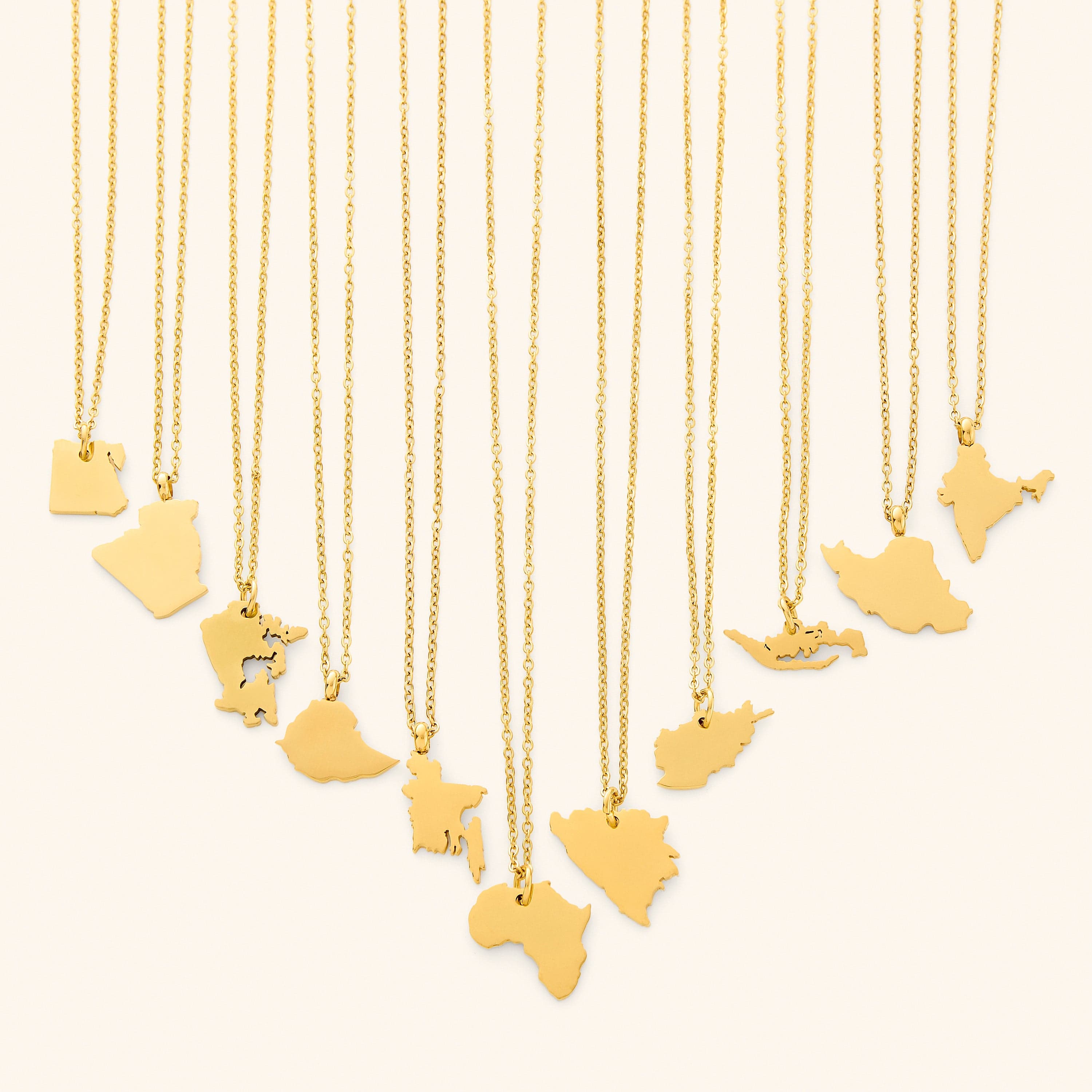 The image shows a collection of delicate gold necklaces hanging vertically against a light beige background. Each necklace features a flat, polished gold pendant shaped like the outline of a different country or region.