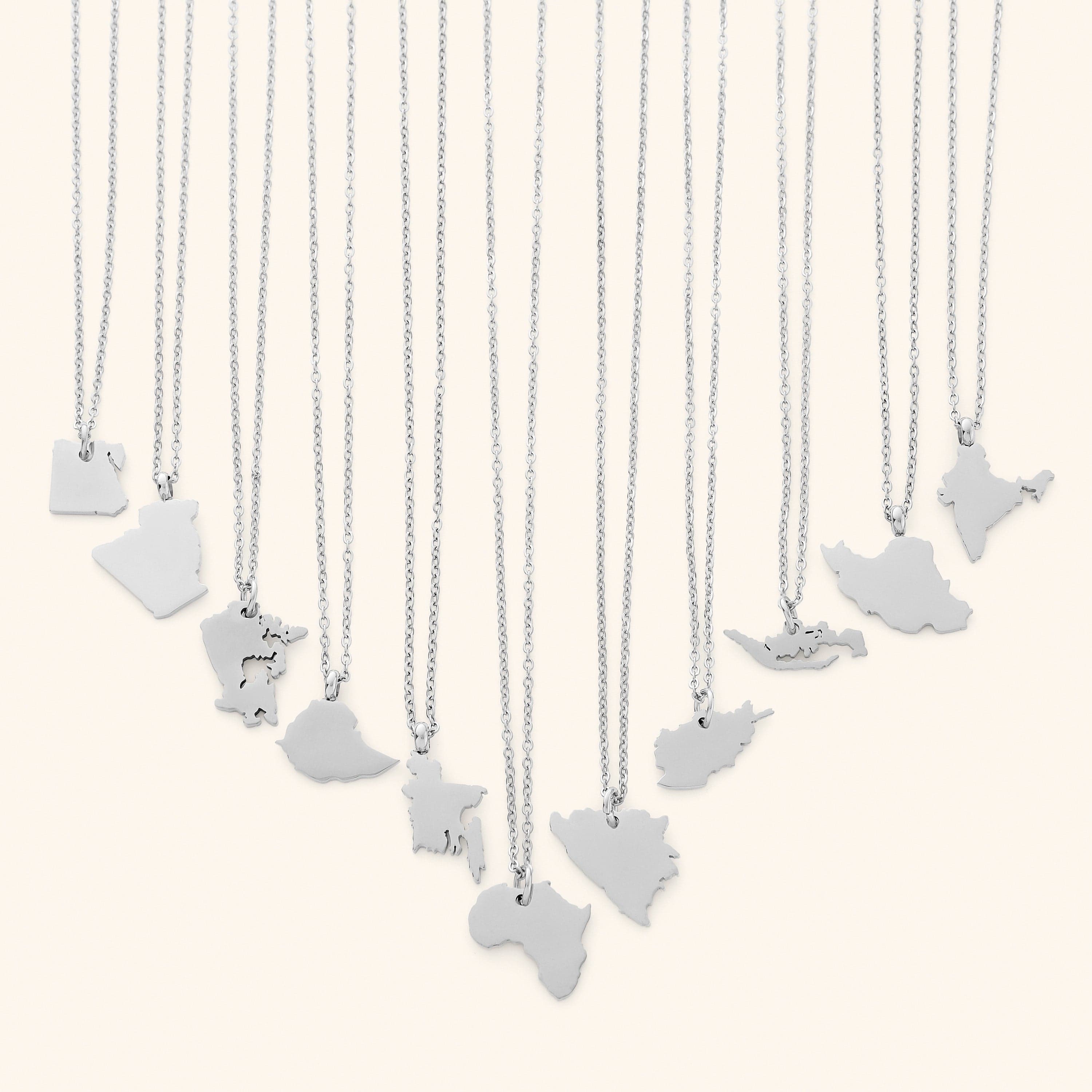 The image shows a collection of delicate silver necklaces hanging vertically against a light beige background. Each necklace features a flat, polished silver pendant shaped like the outline of a different country or region.