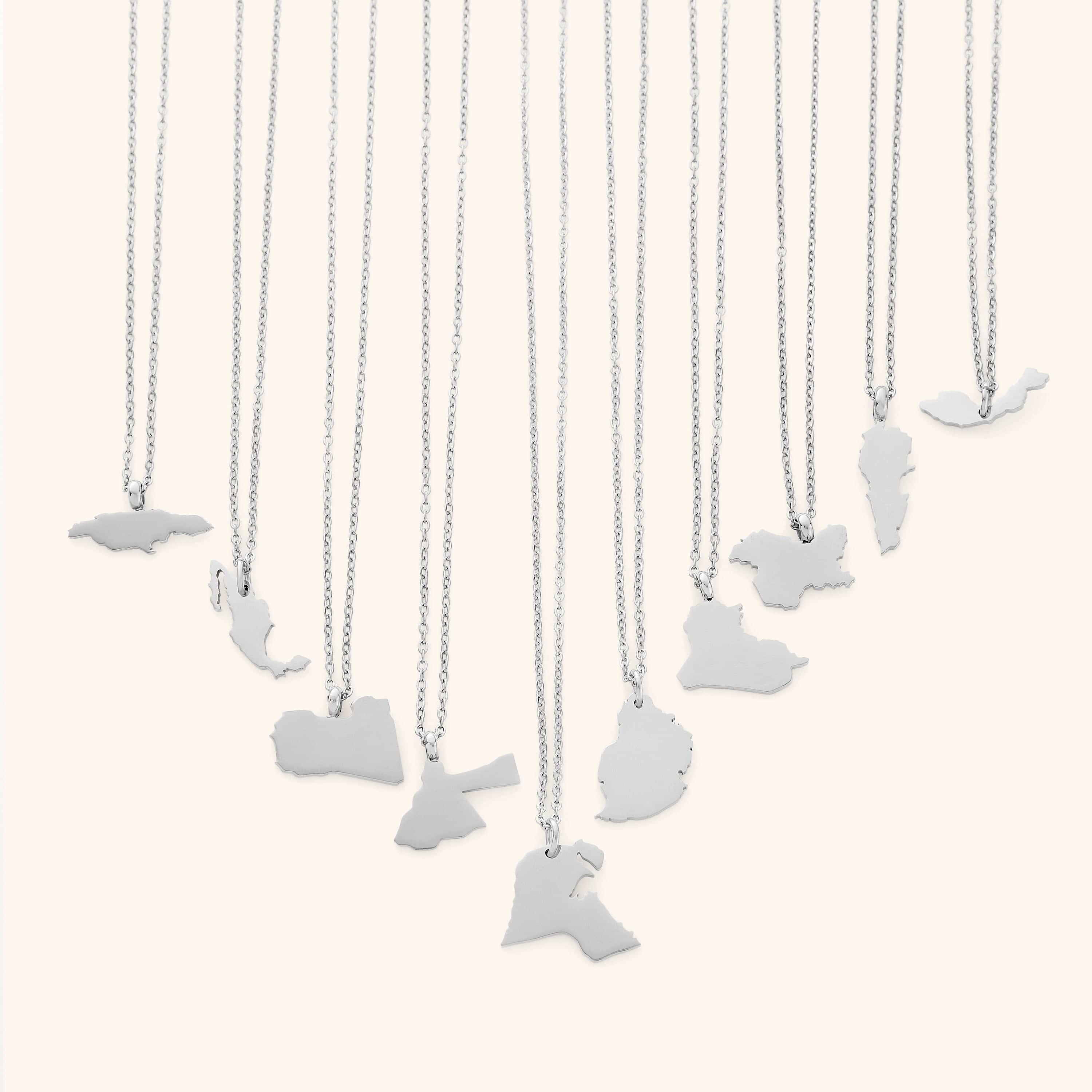 The image shows a collection of delicate silver necklaces hanging vertically against a light beige background. Each necklace features a flat, polished silver pendant shaped like the outline of a different country or region.