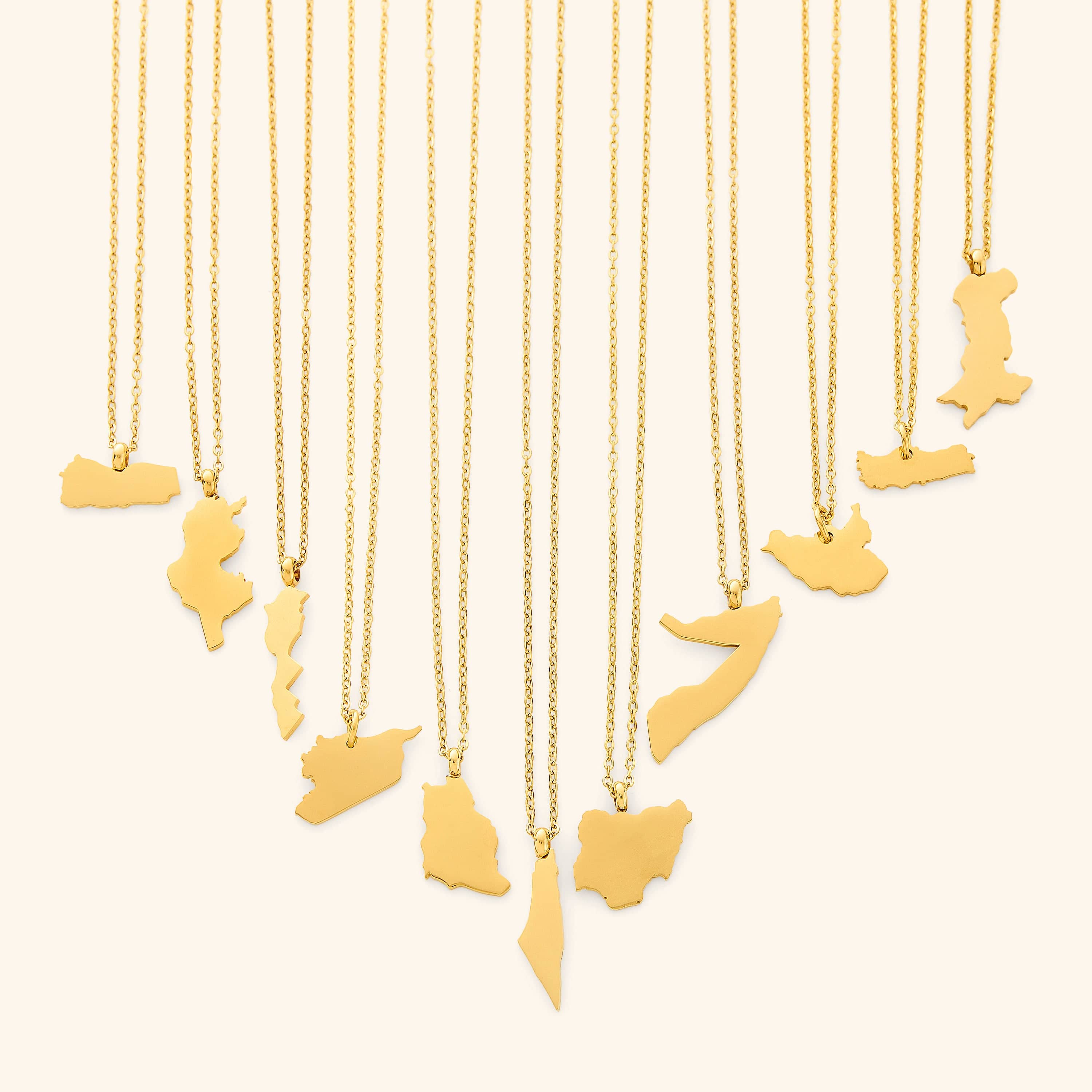 The image shows a collection of delicate gold necklaces hanging vertically against a light beige background. Each necklace features a flat, polished gold pendant shaped like the outline of a different country or region.
