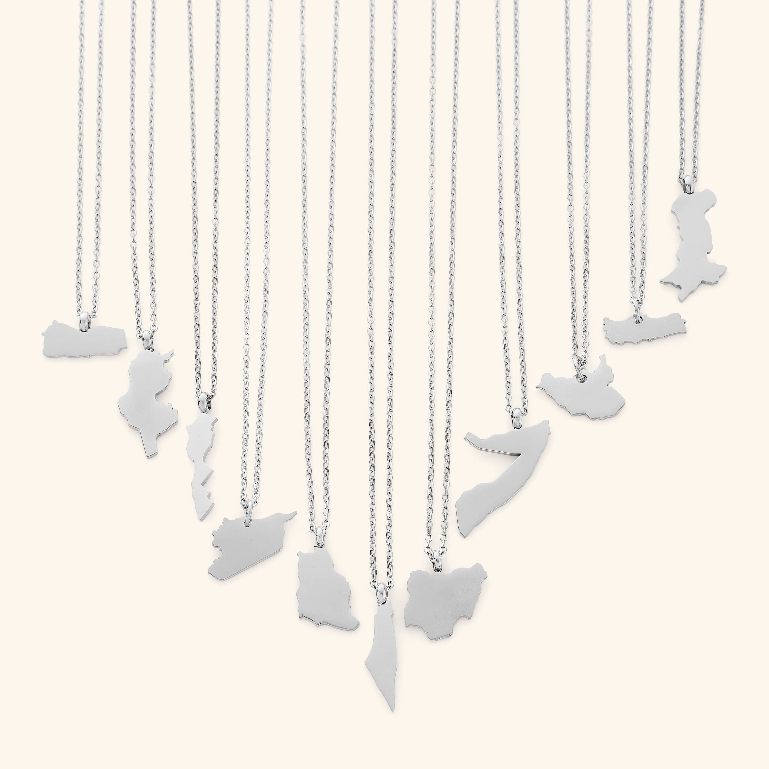 The image shows a collection of delicate silver necklaces hanging vertically against a light beige background. Each necklace features a flat, polished silver pendant shaped like the outline of a different country or region.