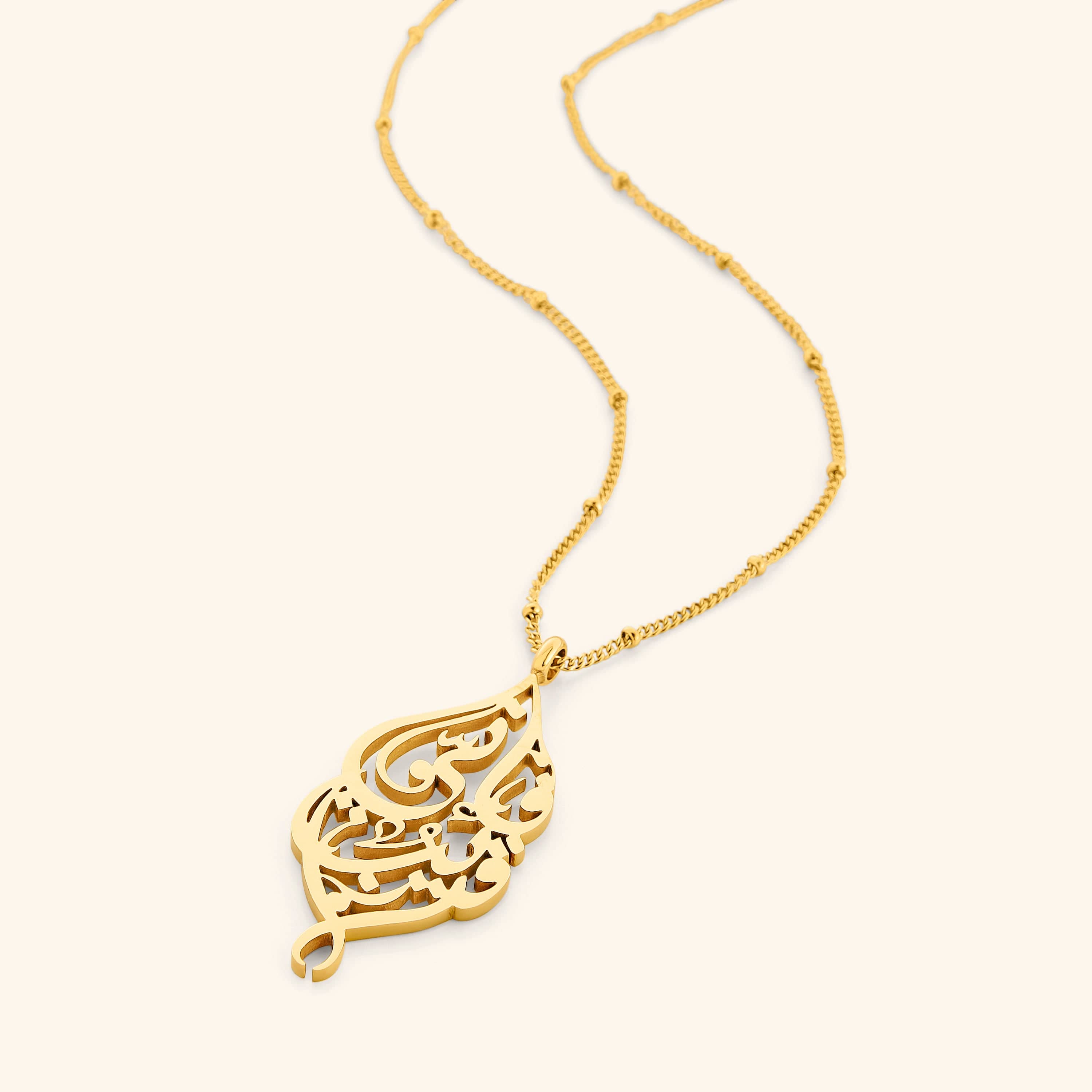 Flat lay of a gold necklace with a pendant that features Arabic Calligraphy of "if My servants ask about Me, indeed I am near" design on an elegant dotted chain.