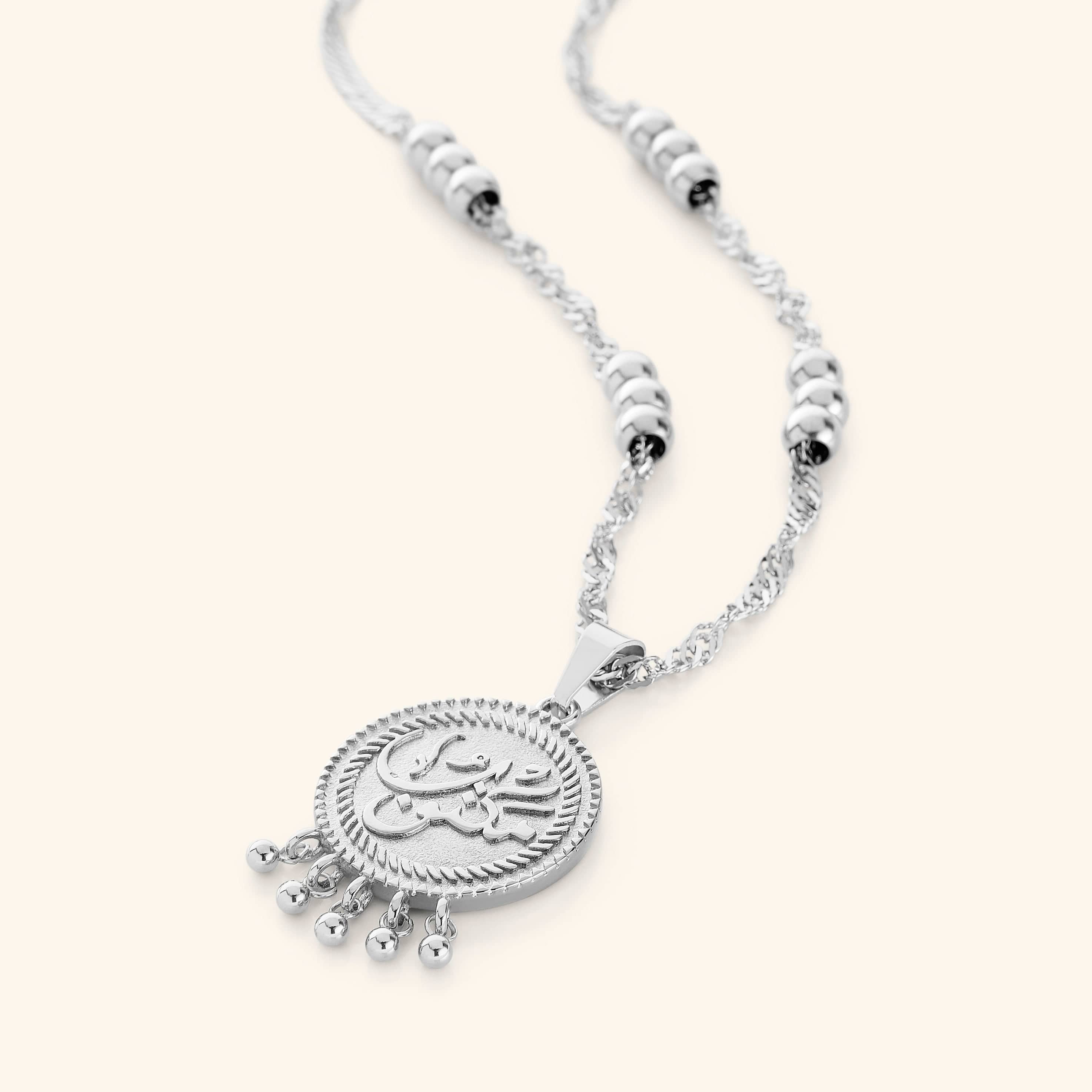 A silver  necklace that features a coin pendant with the inscription: مكتوب—"Maktub", twisted chains, burst orbs, and the delicate dangling dots add movement and elegance, 