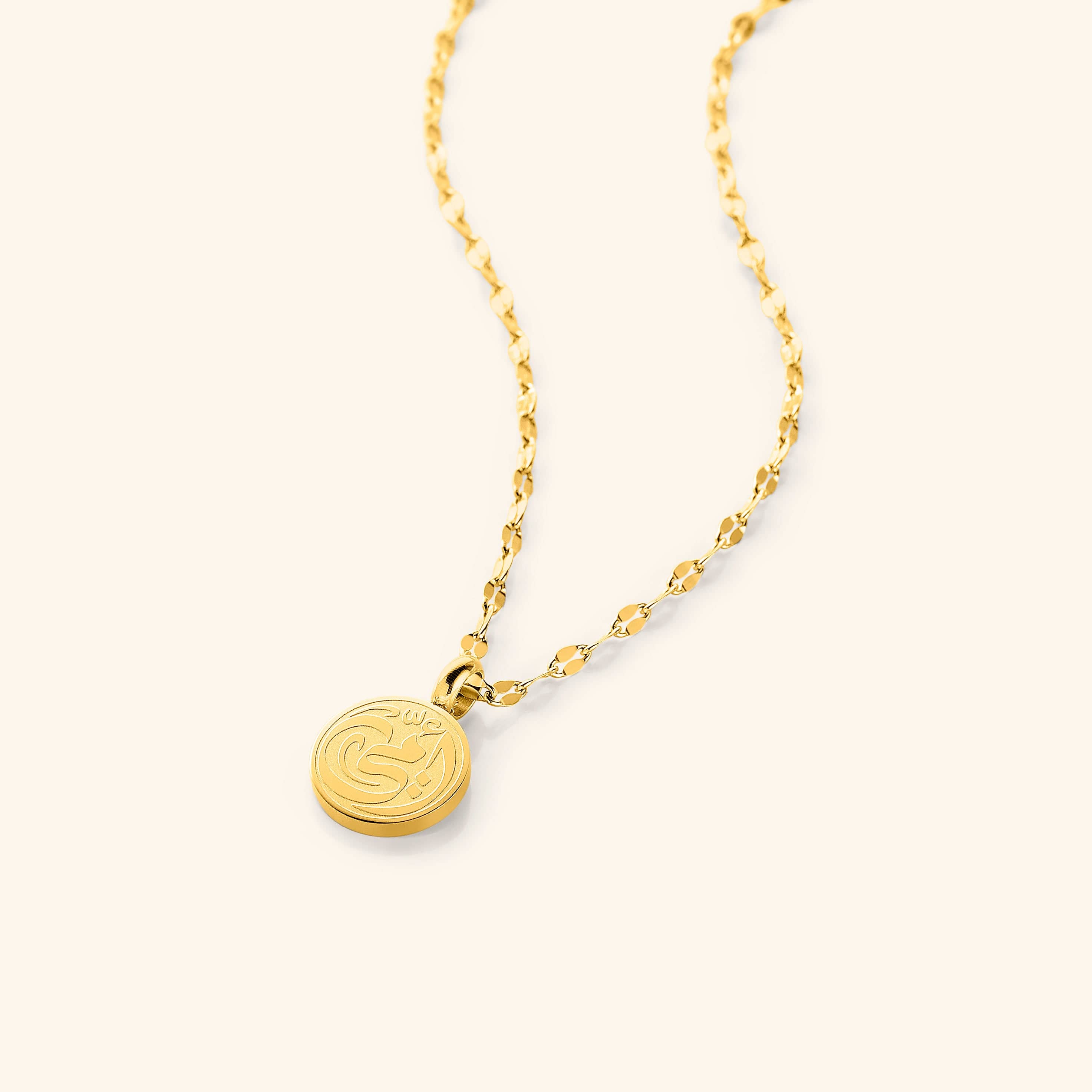 A close-up of the My Mother Necklace in 18K gold plating. The round pendant features the Arabic word "أمي" ("Ummi" – My Mother) engraved in elegant calligraphy, suspended from a delicate gold Flutter Chain.