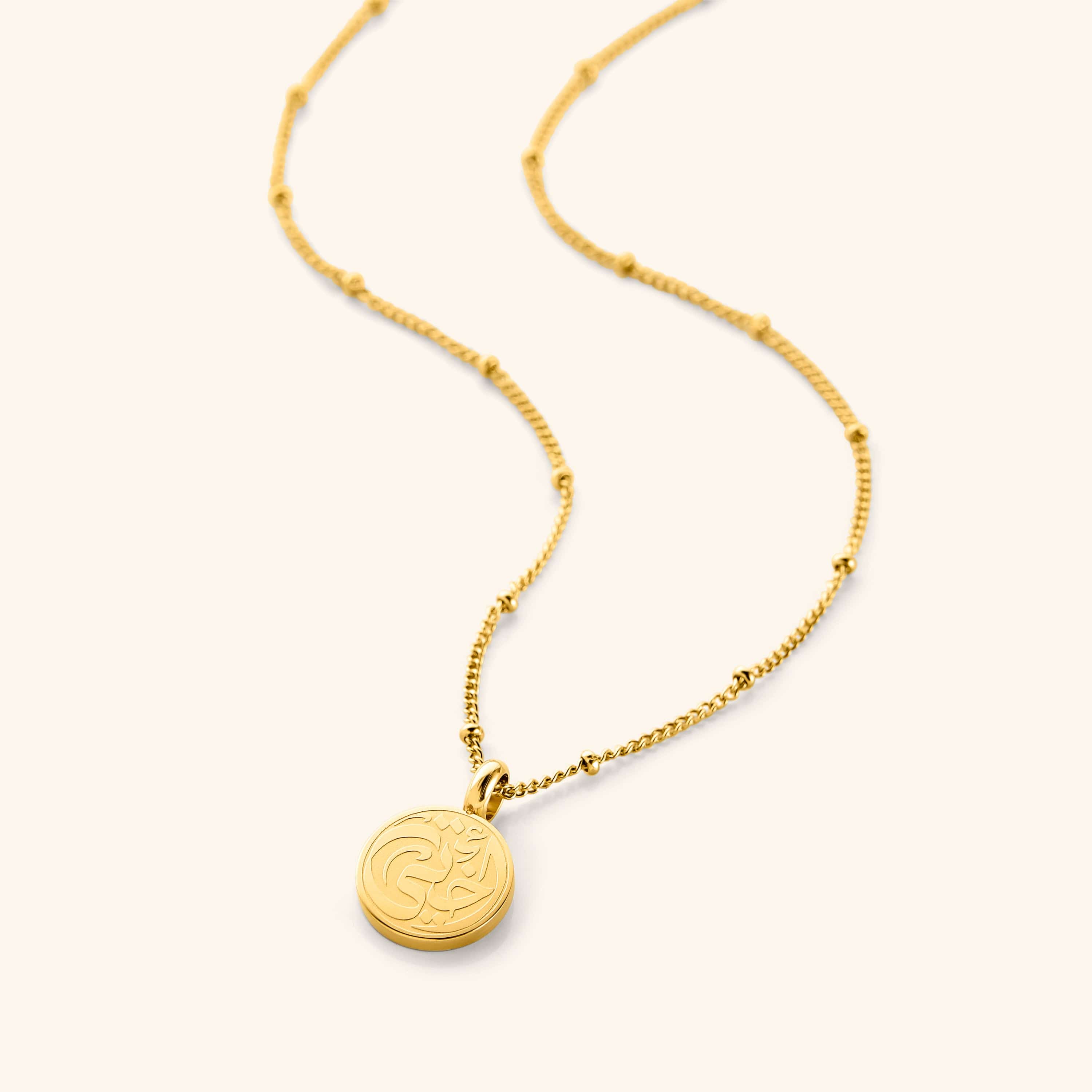 Flat lay o the gold My Sister Necklace against a white background. Sister Necklace features "my sister" inscribed in Arabic (أختي) on a dainty token, finished with a dotted chain.