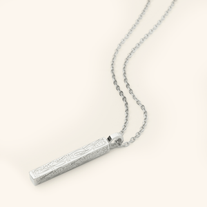 Photo of a silver necklace that features the Arabic Calligraphy of  'with every hardship comes ease' that is engraved intricately all along this timeless bar pendant. 