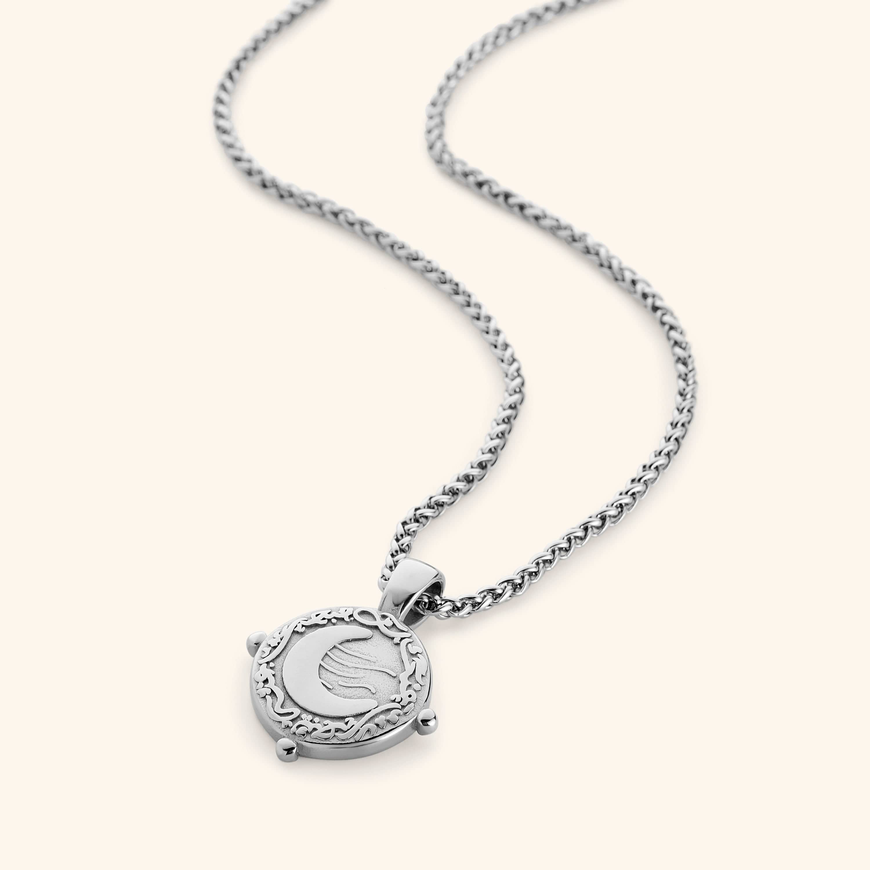 Silver necklace with a round pendant featuring the engraved phrase 'What you seek is seeking you' in Arabic calligraphy. The design includes a raised crescent moon and a flowing motif, framed with vine detailing and three small bead accents around the border. 
