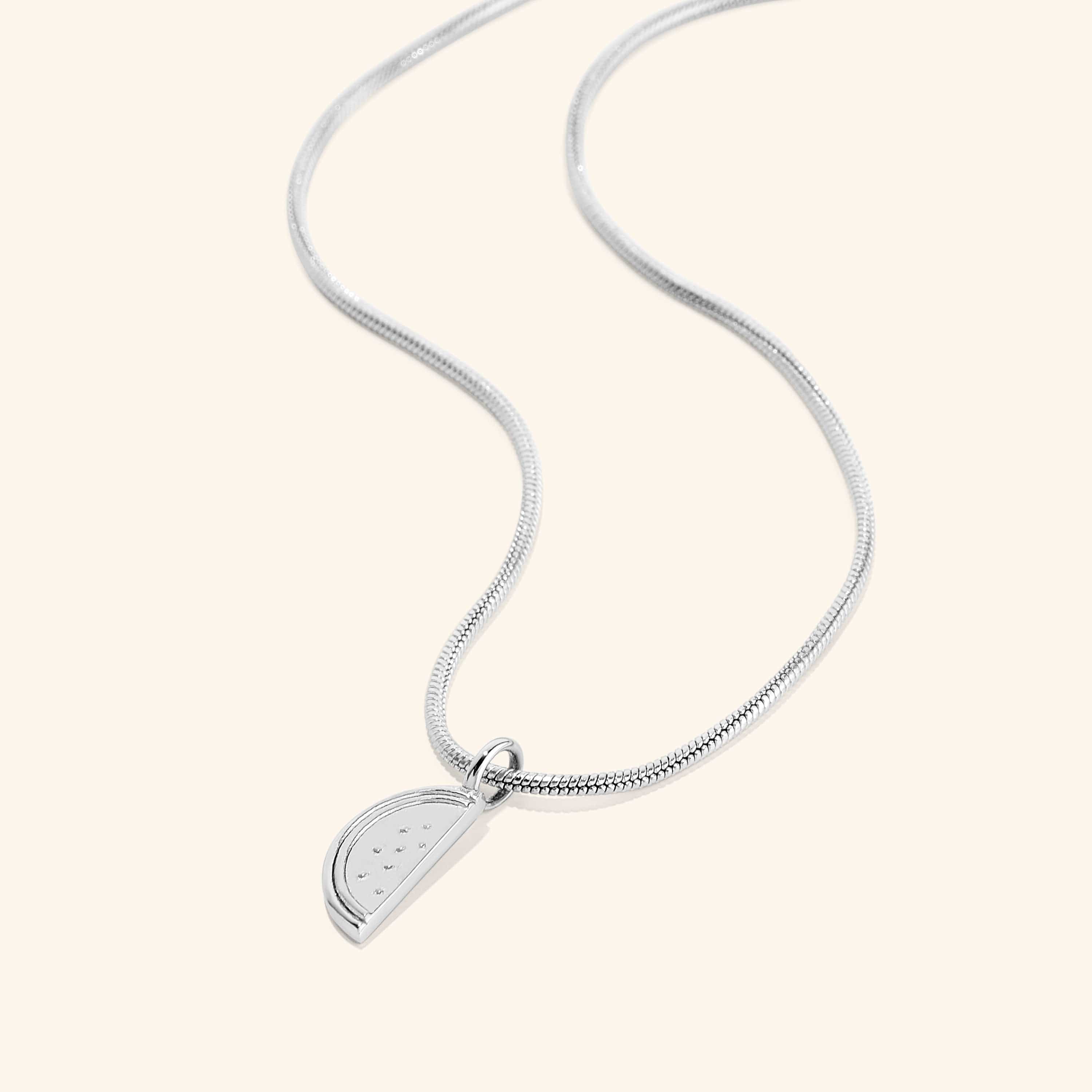 A silver necklace with a pendant shaped like a slice of a watermelon. The watermelon pendant has small indentations representing seeds. The necklace features a thin, flexible silver snake chain. The necklace is displayed on a light beige background.