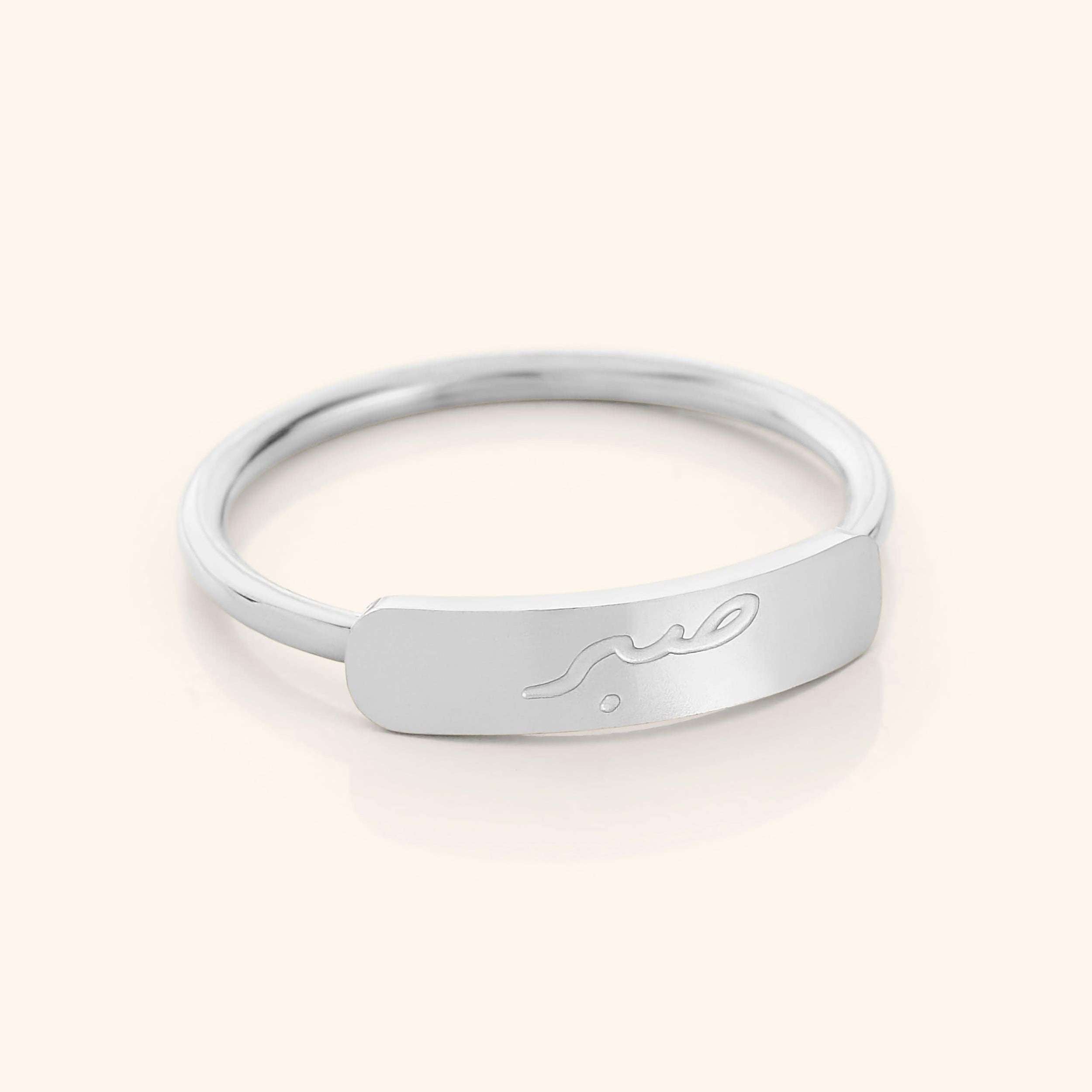 Slim silver ring with a rectangular bar centerpiece engraved with the Arabic word meaning "patience," photographed on a neutral background.