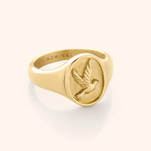 Close-up of a gold signet-style ring with a raised circular face engraved with a flying dove, photographed on a neutral background. The inside of the band features the engraved word "NOMINAL."
