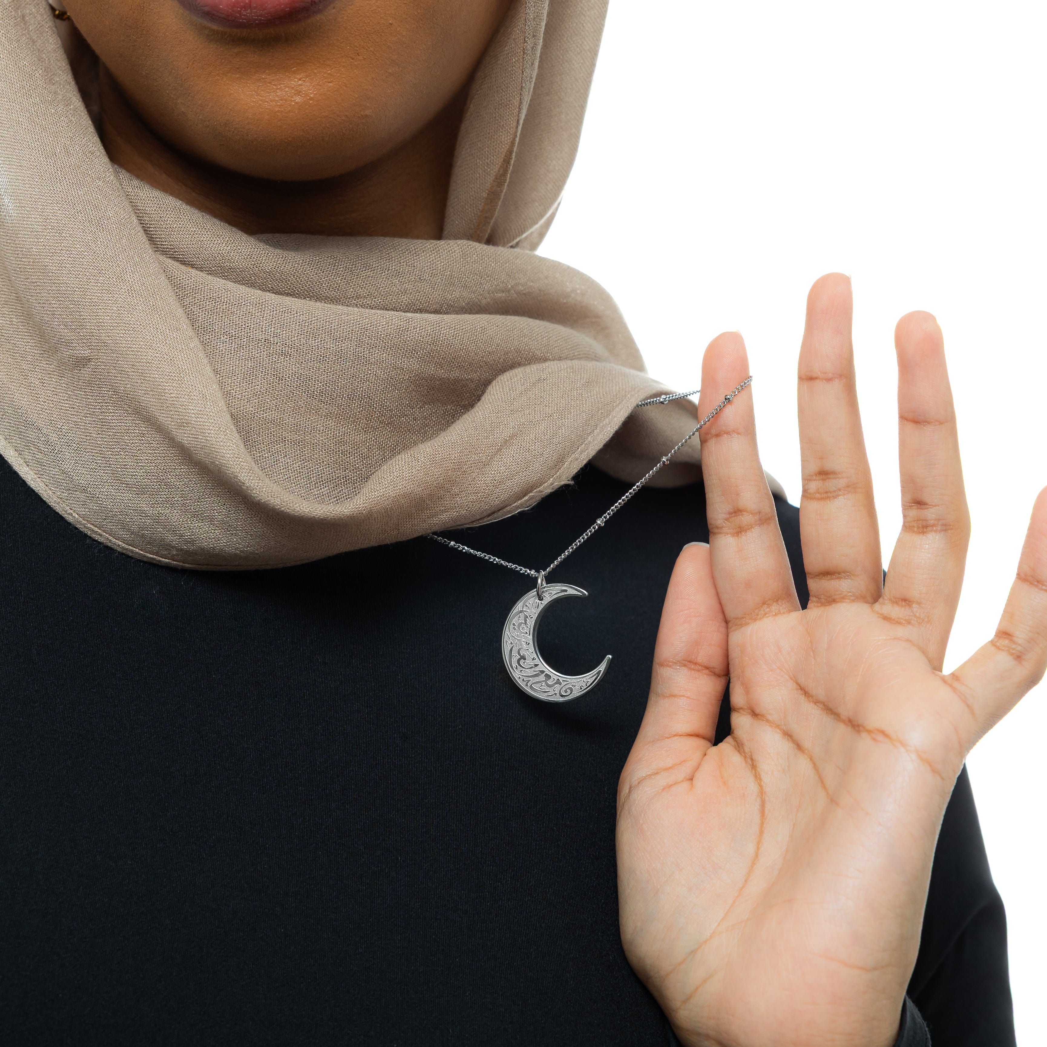 A close-up of a silver necklace with a crescent moon pendant. The pendant is inscribed with Arabic script. The necklace is held up against a blurred background of a person wearing a black top. The focus is on the detail of the pendant and the script. Given the inscription "With Hardship Comes Ease" and the crescent moon, which holds significance in Islamic culture, it's plausible that the script is related to Islamic teachings and the necklace carries religious or inspirational meaning for the wearer.