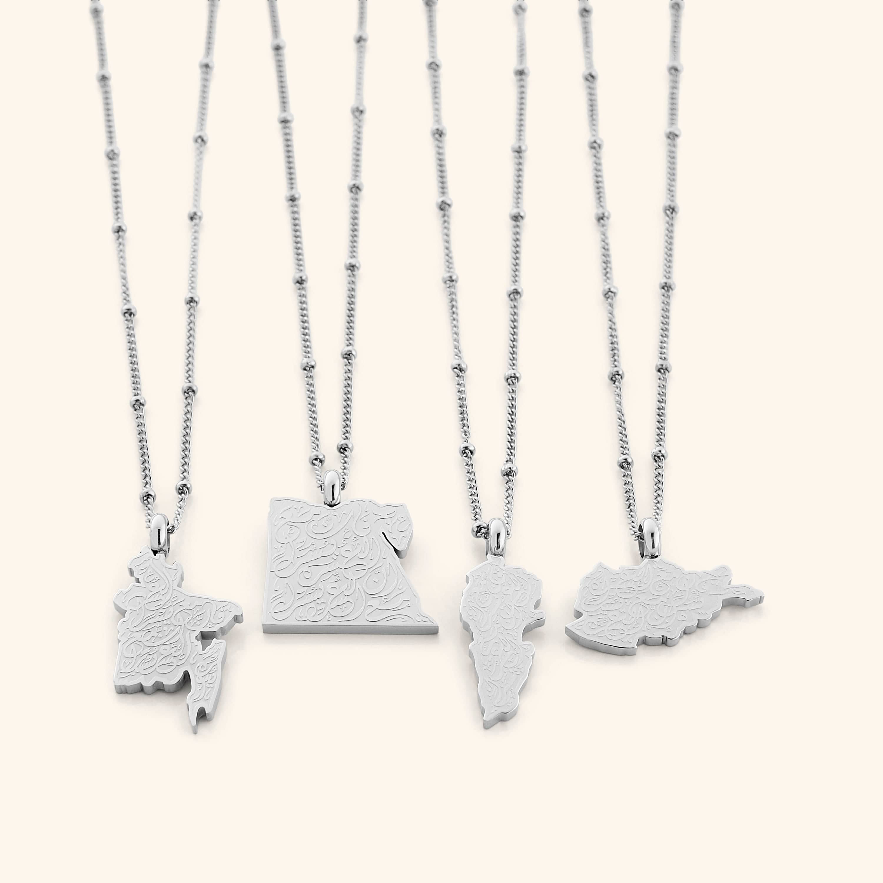Four silver "With Hardship Comes Ease" Map Necklaces arranged side by side on a white background, each pendant shaped like a different map and engraved with Arabic calligraphy, showcasing subtle differences in map outlines and silver chain styles.