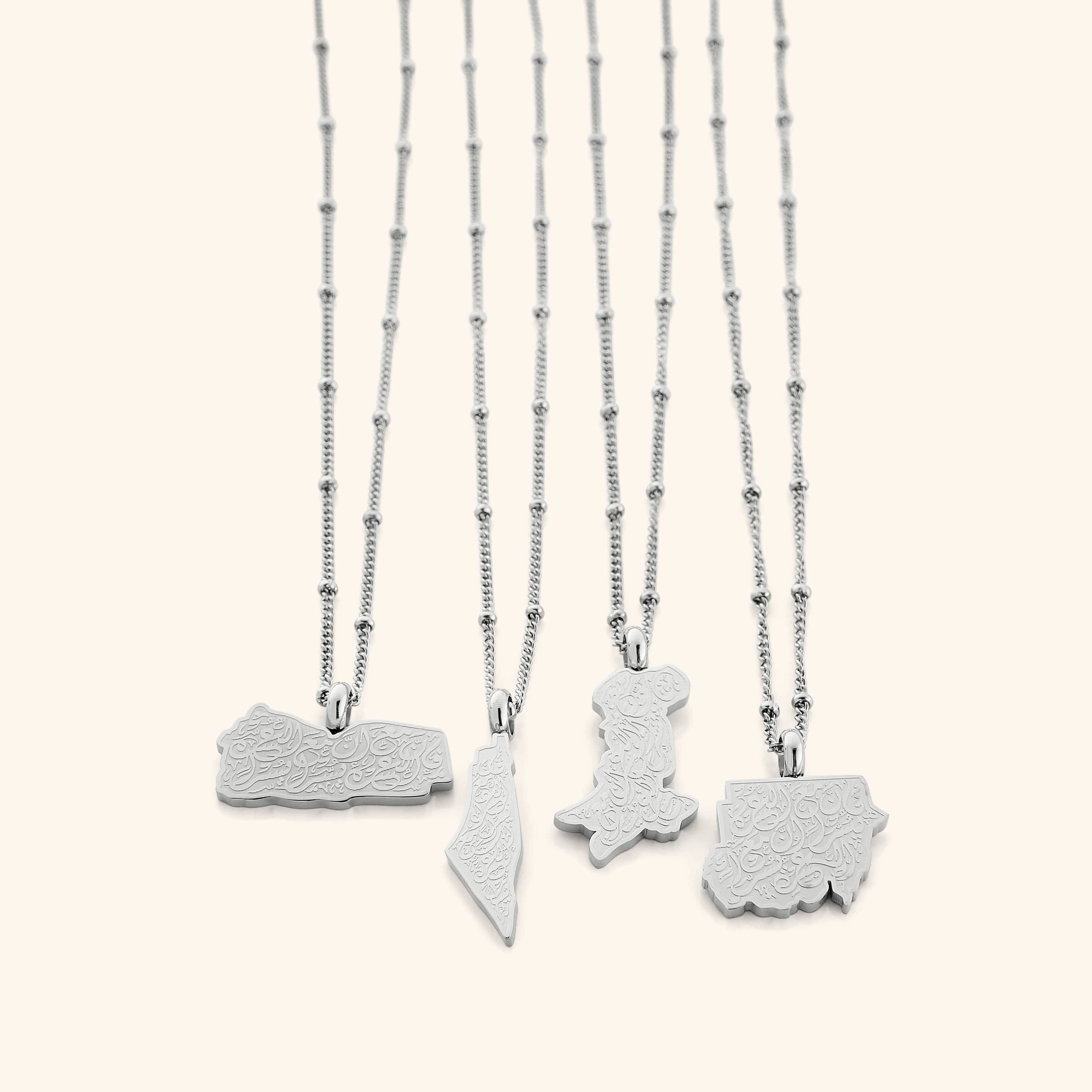 Four silver "With Hardship Comes Ease" Map Necklaces arranged side by side on a white background, each pendant shaped like a different map and engraved with Arabic calligraphy, showcasing subtle differences in map outlines and silver chain styles.